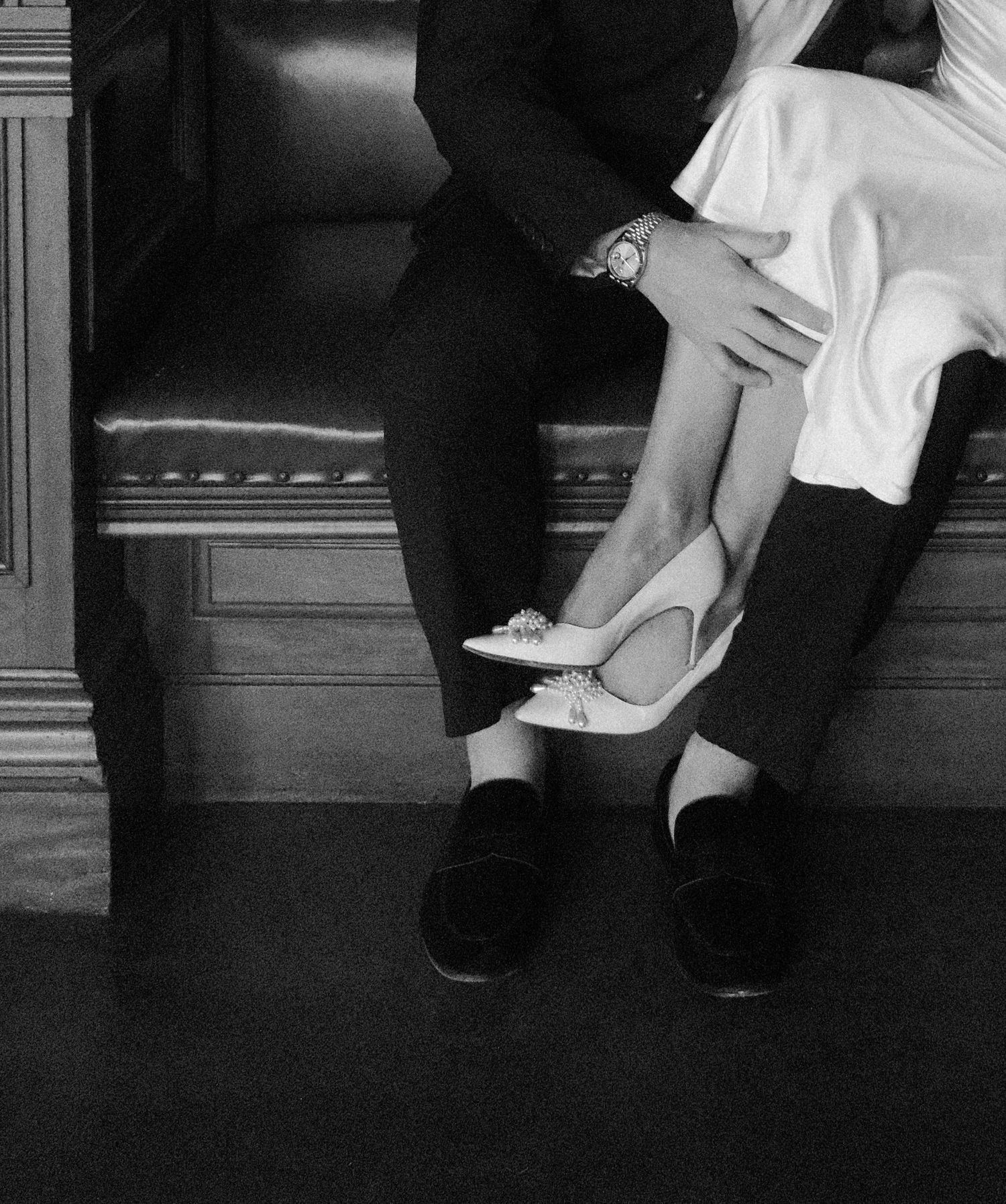 Close-up of bride in white heels and groom in black loafers sitting together on a bench.film-inspired
