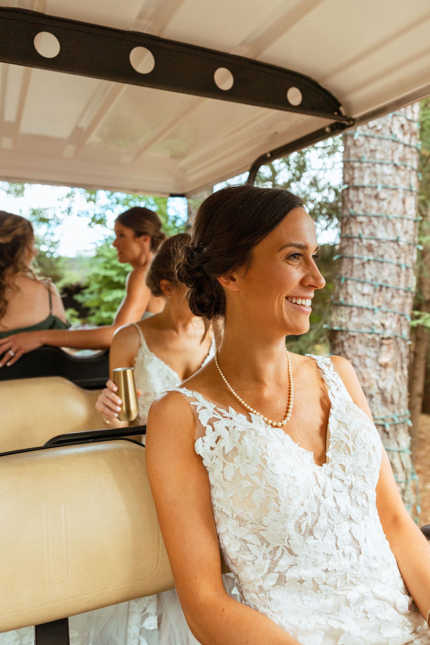 Smiling bride in lace gown and pearl necklace rides golf cart holding champagne on wedding day.