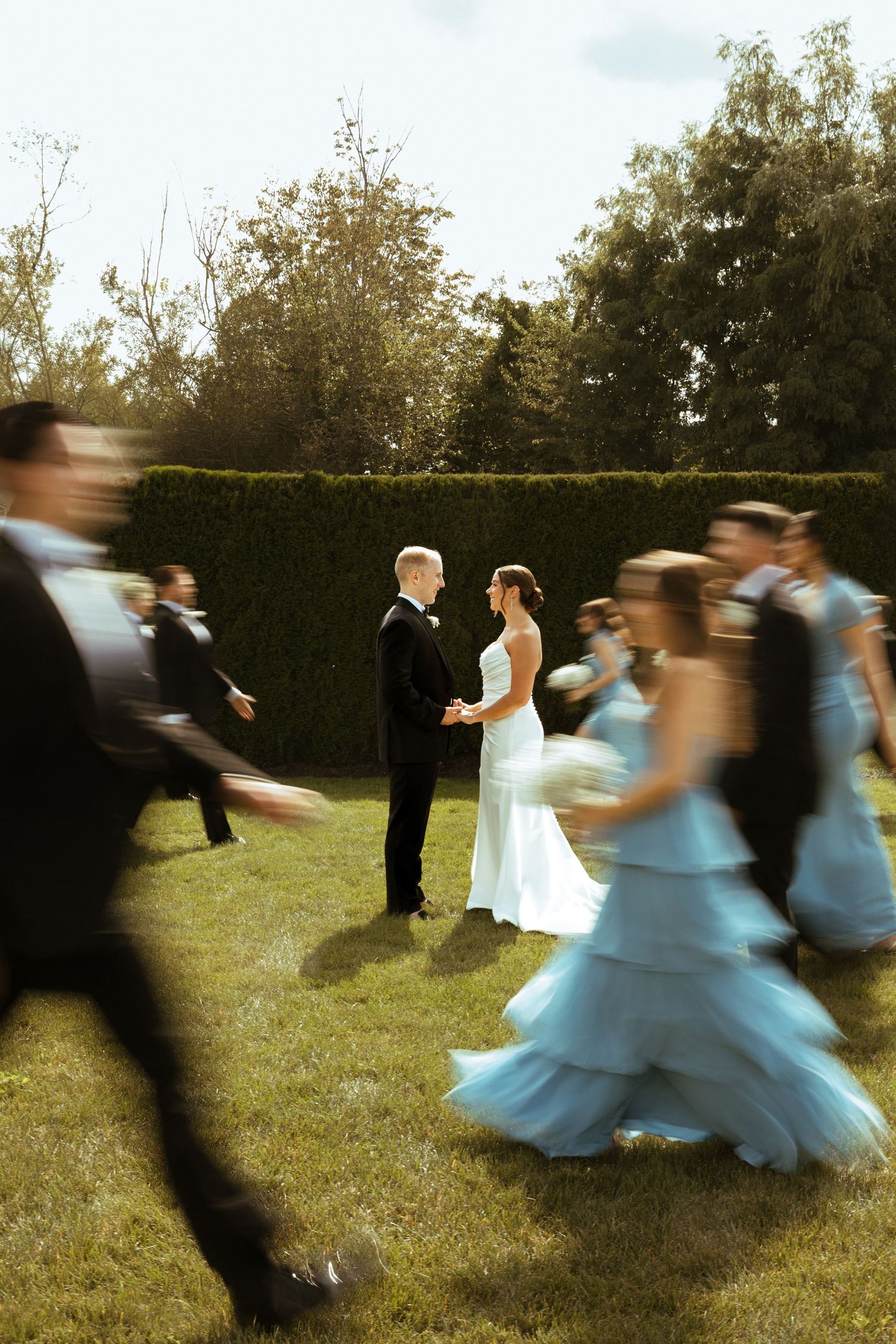 Bride and groom stand still as wedding party in blue gowns and black suits swirl around them outdoors.