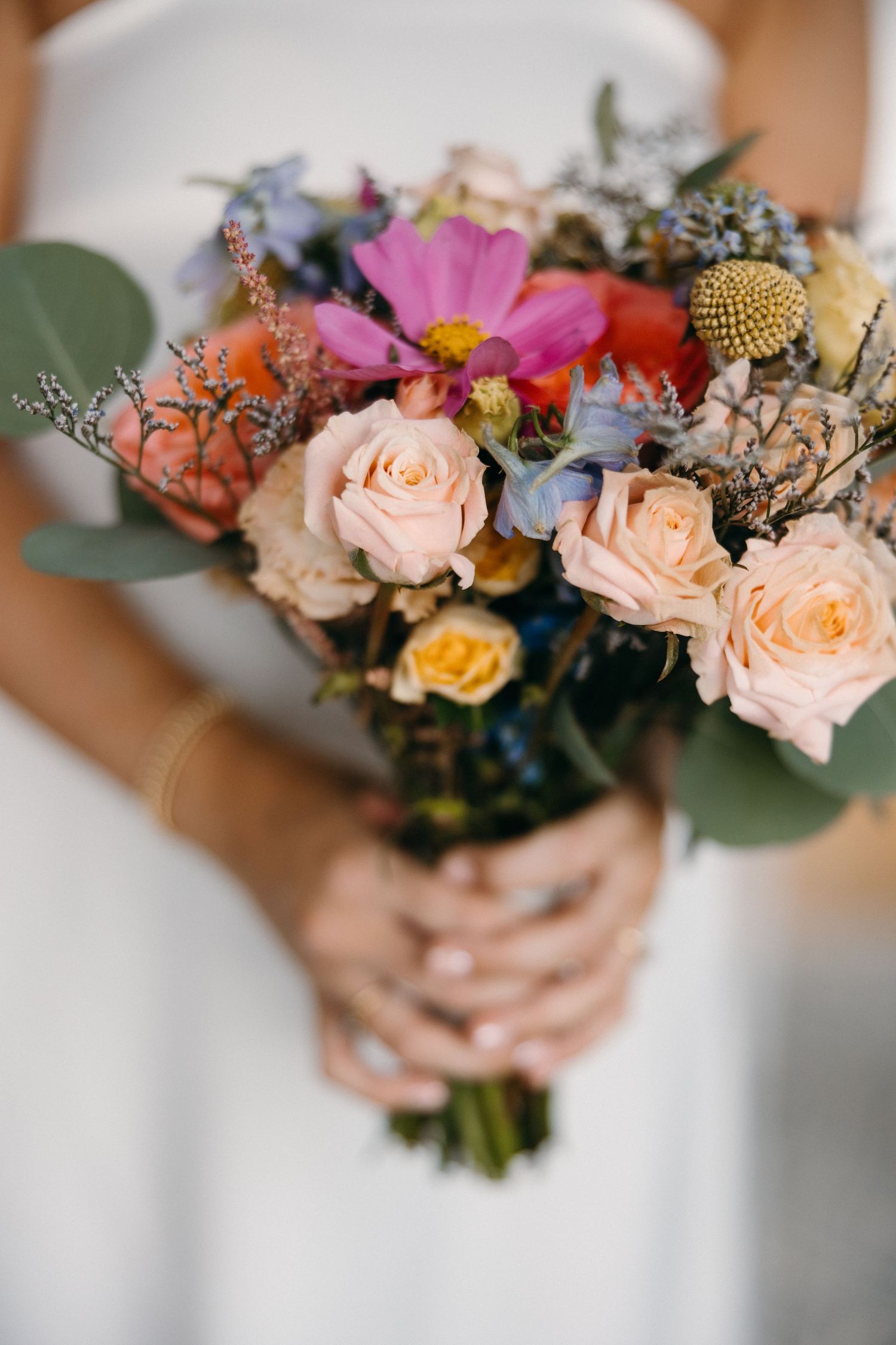 Bride holding colorful wedding bouquet with roses, wildflowers, and eucalyptus in white dress.