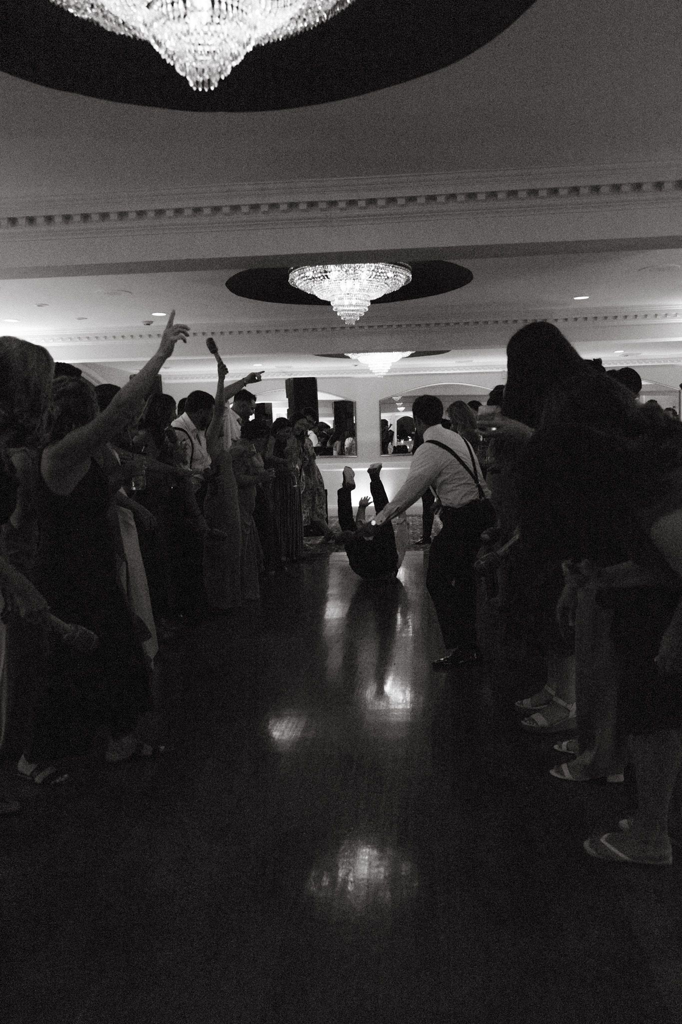 Black and white photo of wedding guests forming a dance circle at elegant ballroom reception with chandeliers.