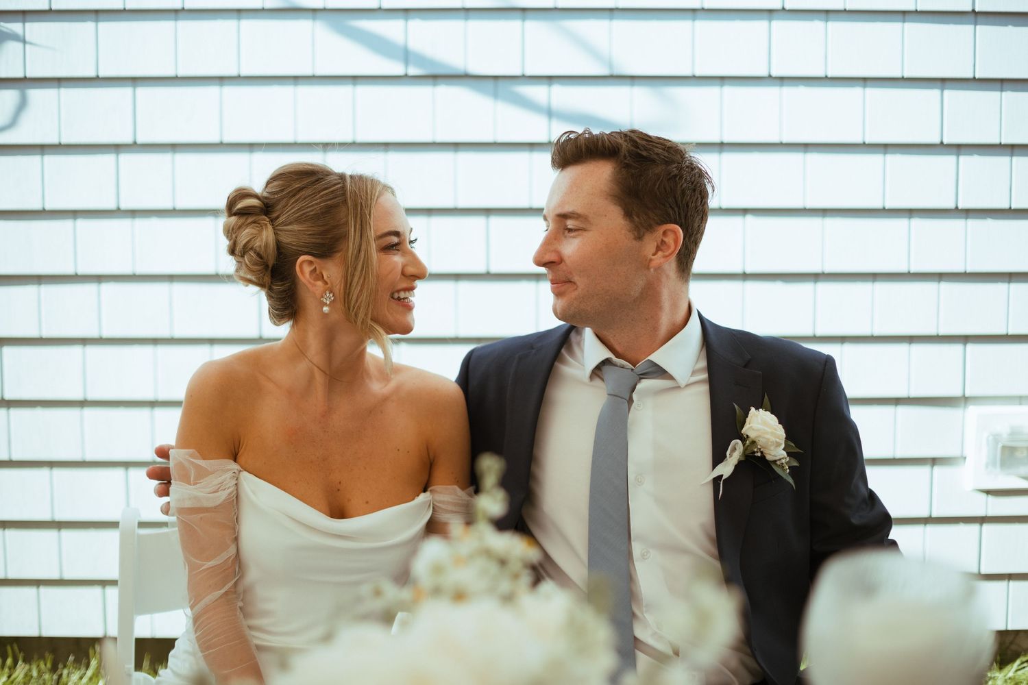 Bride and groom smiling at each other seated at wedding reception table against shingle wall.