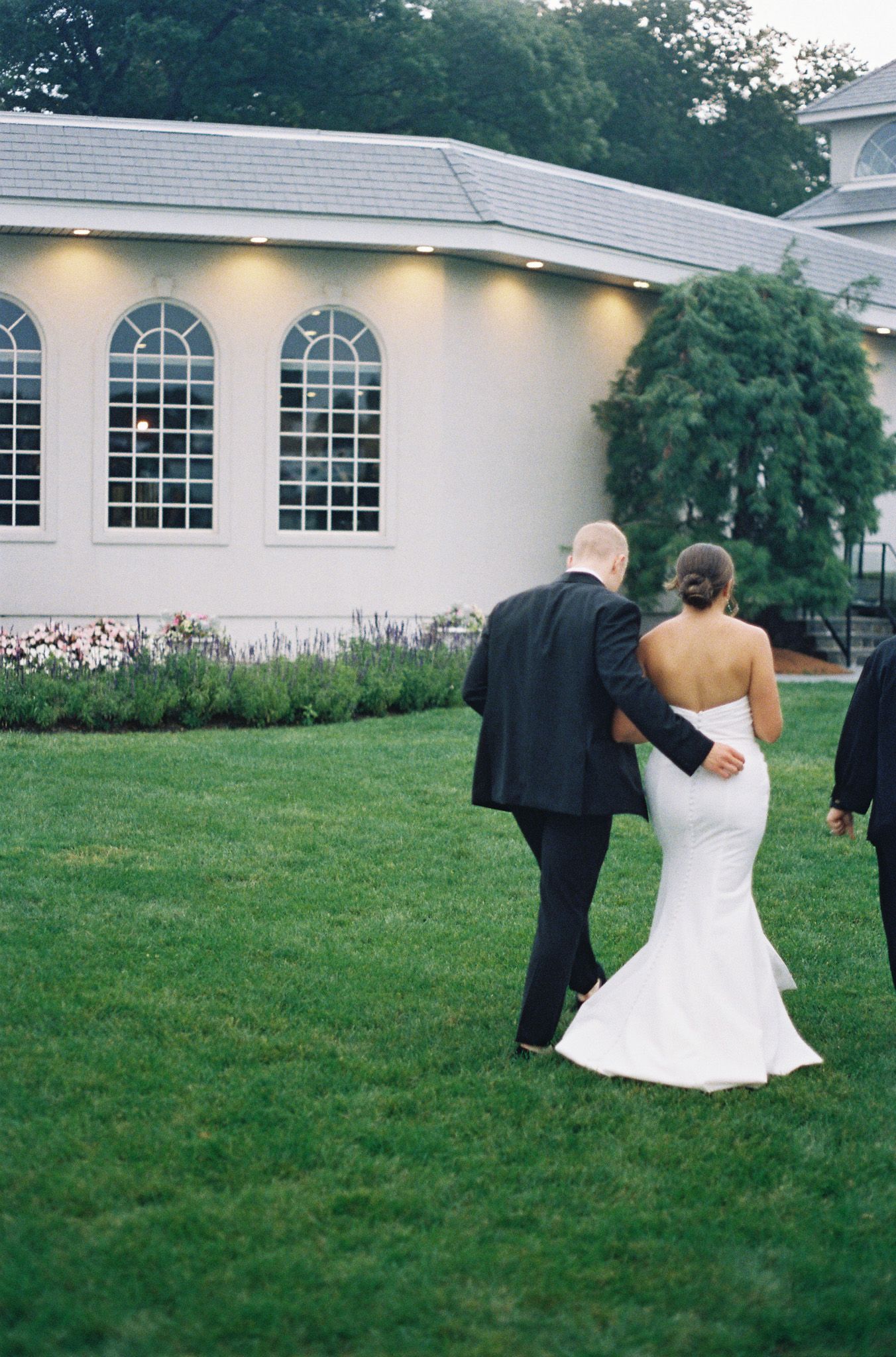 Bride in strapless gown walking with escort across green lawn at elegant venue.