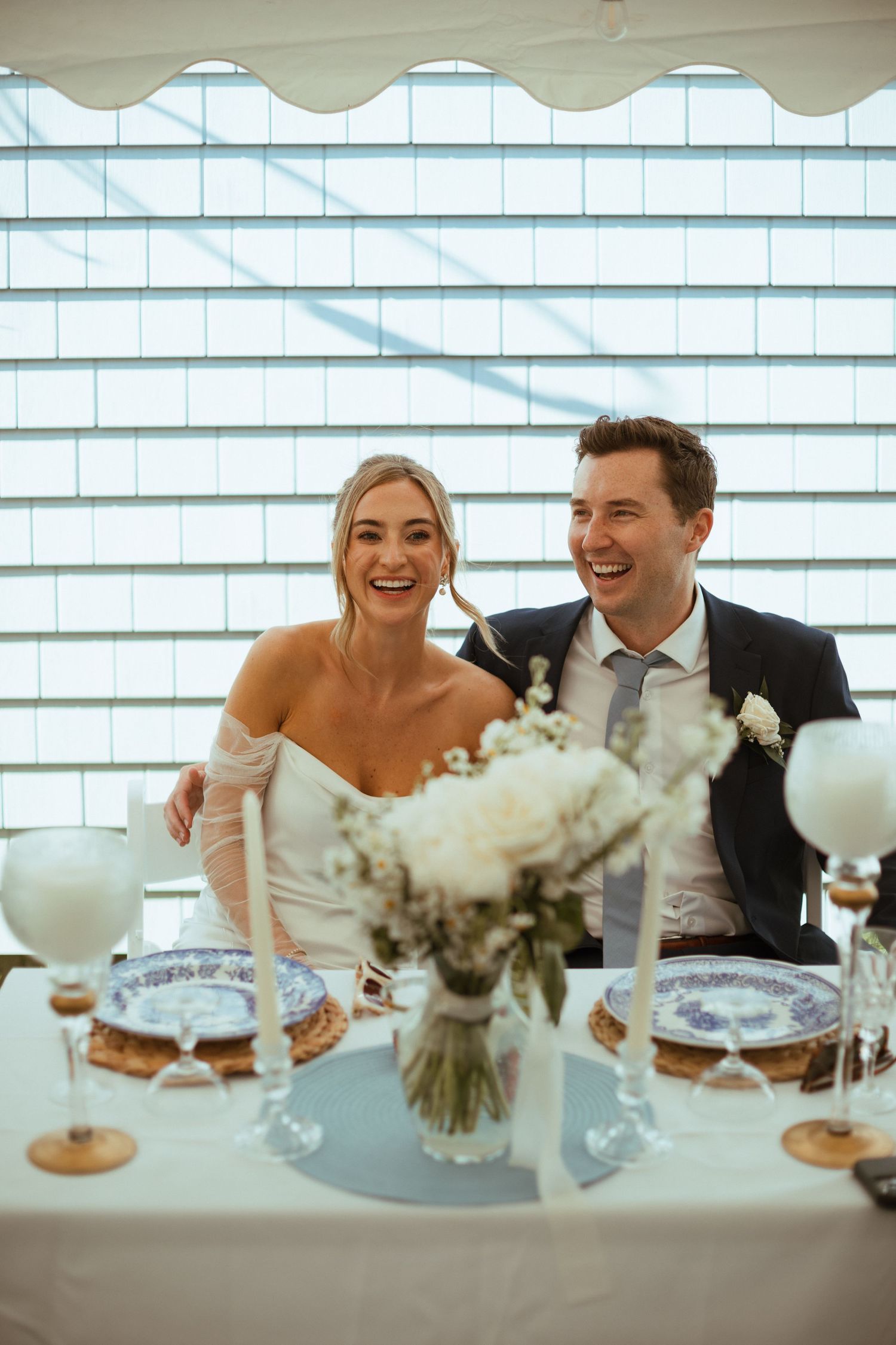 Laughing bride and groom seated at sweetheart table with white floral centerpiece during wedding reception.