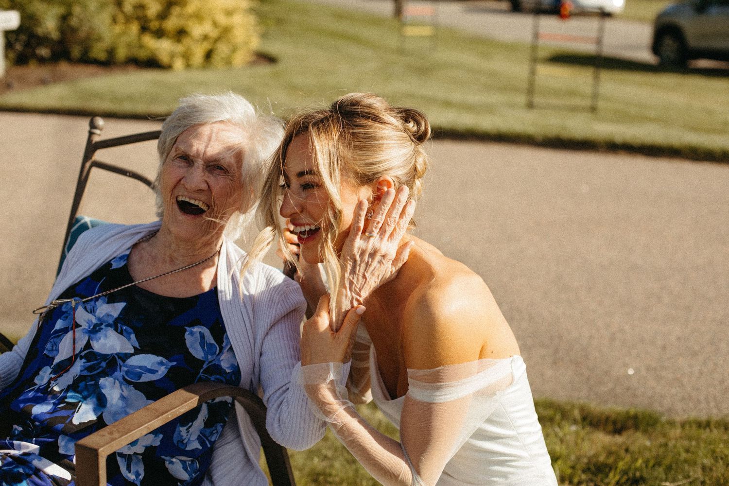 Bride laughing with elderly grandmother in wheelchair during outdoor wedding celebration.