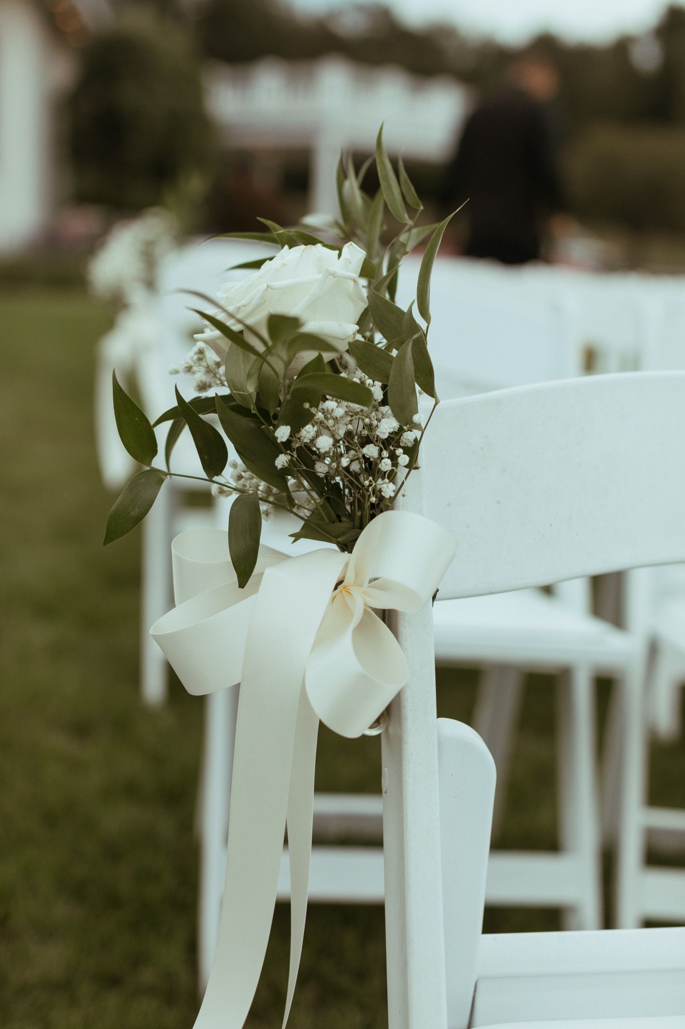 White floral bouquet with ribbon tied to outdoor wedding ceremony chair aisle decoration.