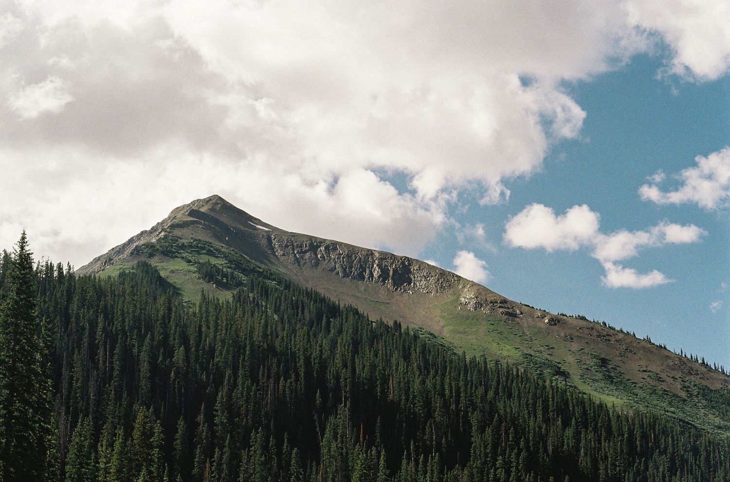 A rocky mountain peak rises above dense evergreen forest under a partly cloudy blue sky.