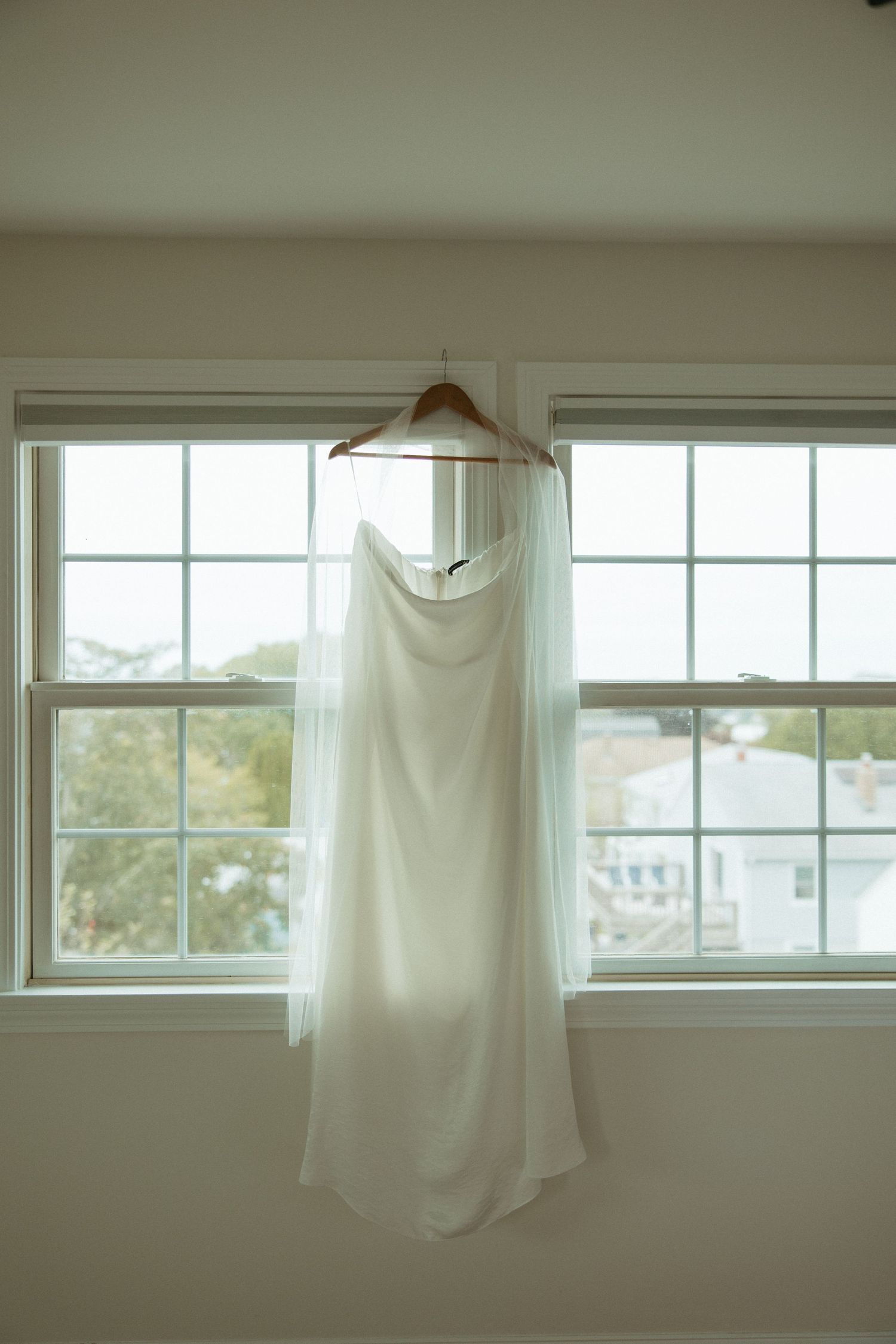 Elegant white wedding dress hanging on wooden hanger in front of window.