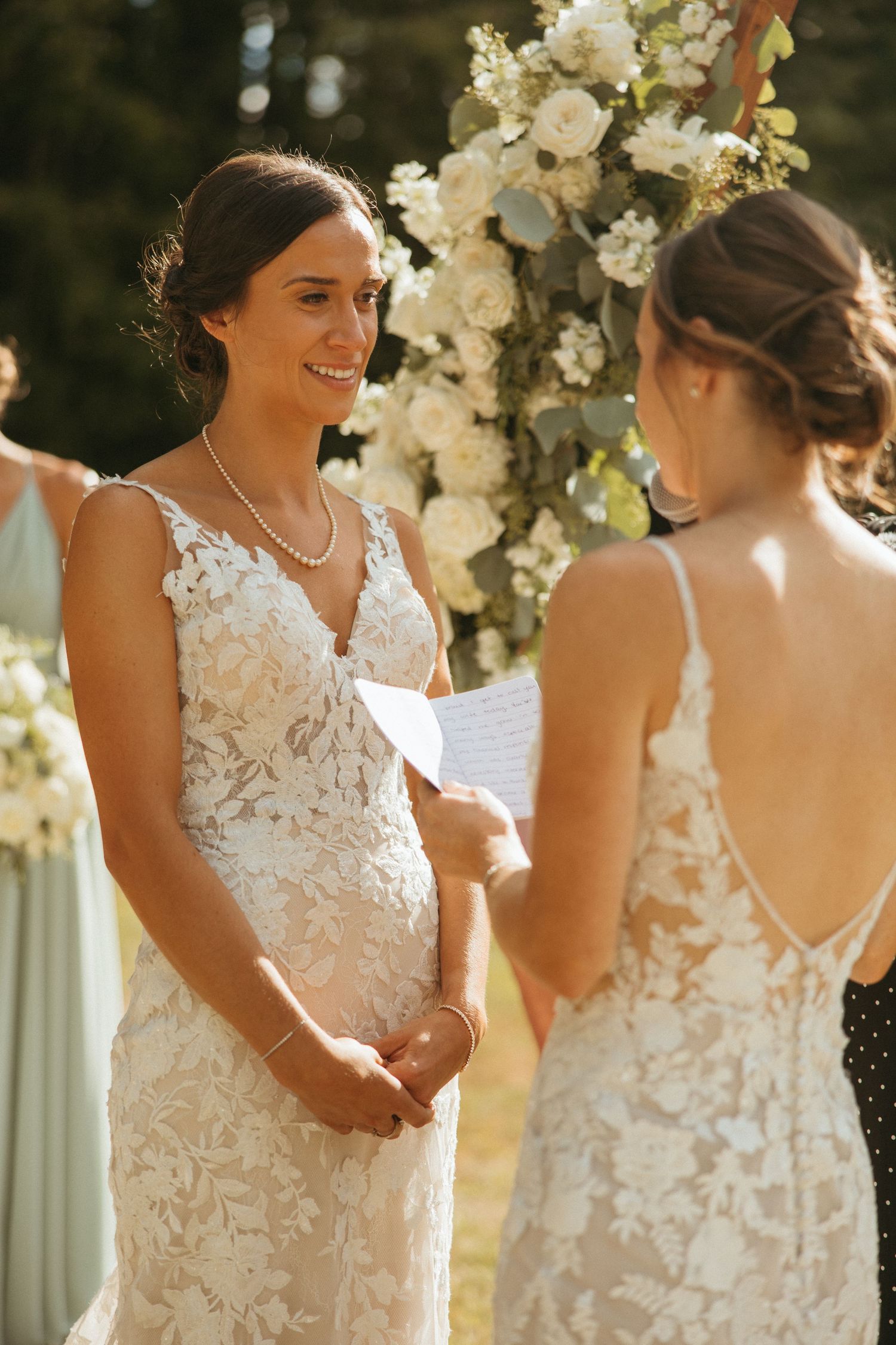 Smiling bride in lace gown listens to vows at outdoor wedding ceremony beneath floral arch.
