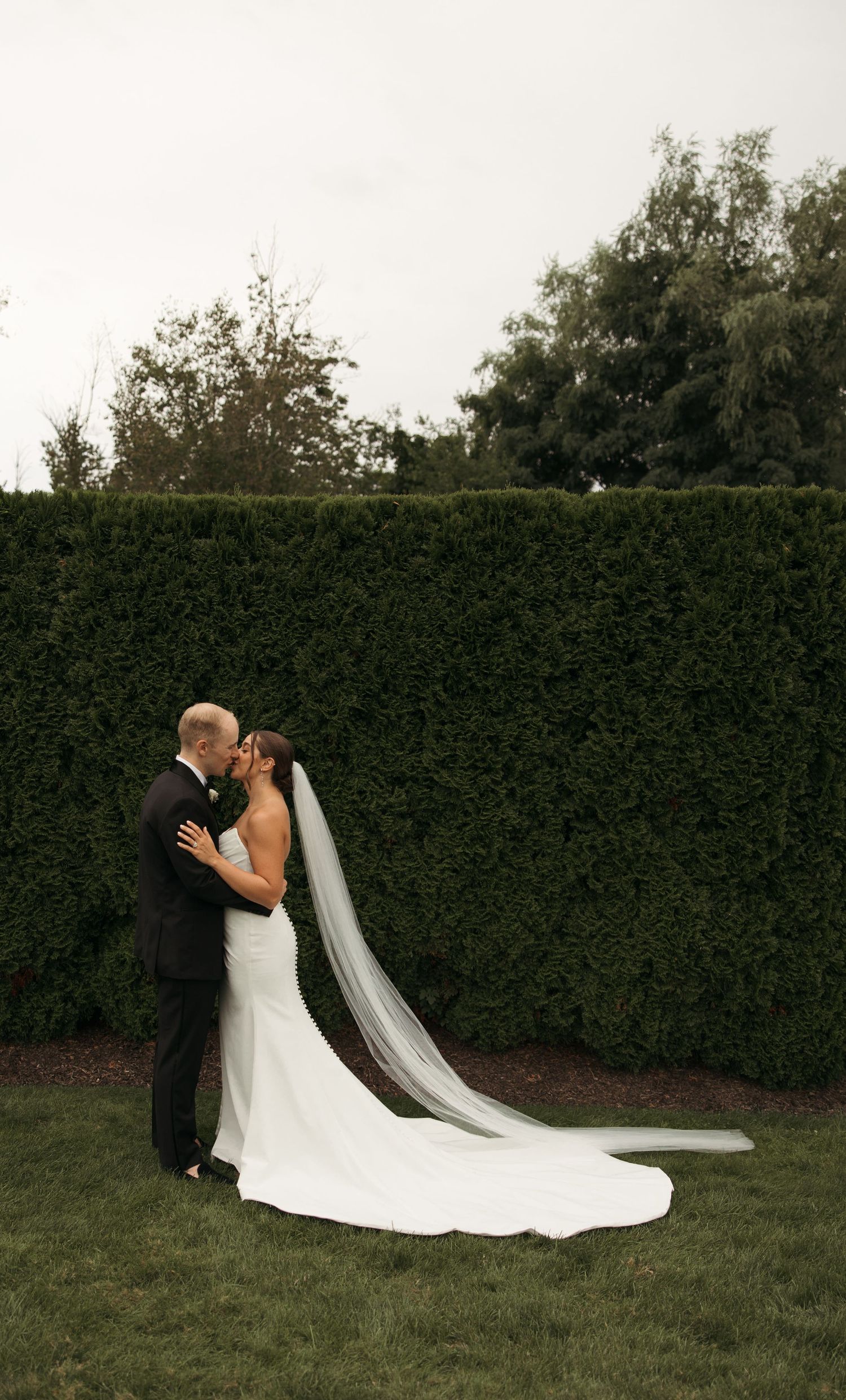 Bride and groom share a kiss on grass lawn with long cathedral veil trailing behind elegant white gown.