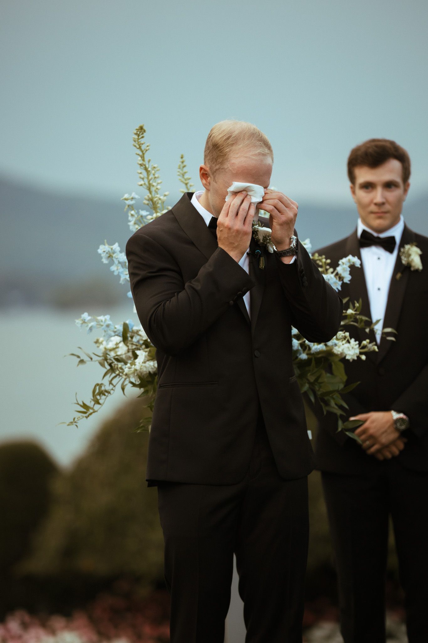 Emotional groom wipes tears with handkerchief at outdoor wedding altar with floral arrangements.