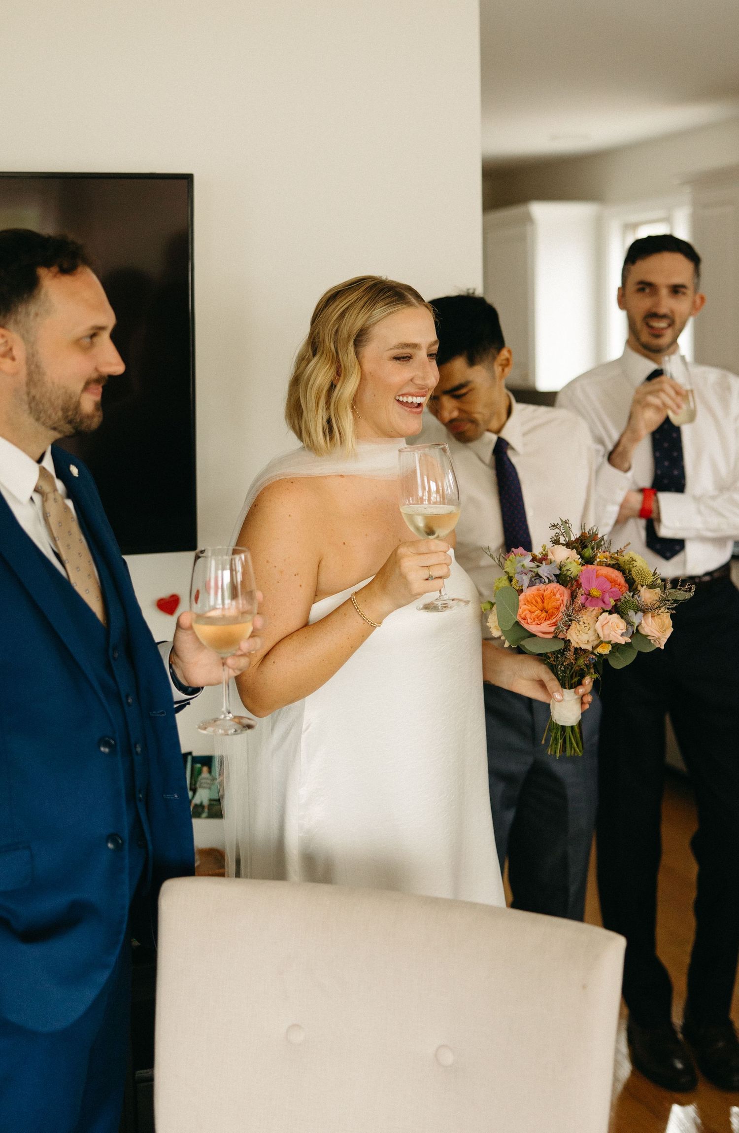 Laughing bride in white dress toasts with wine glass as groom and guests celebrate indoors.