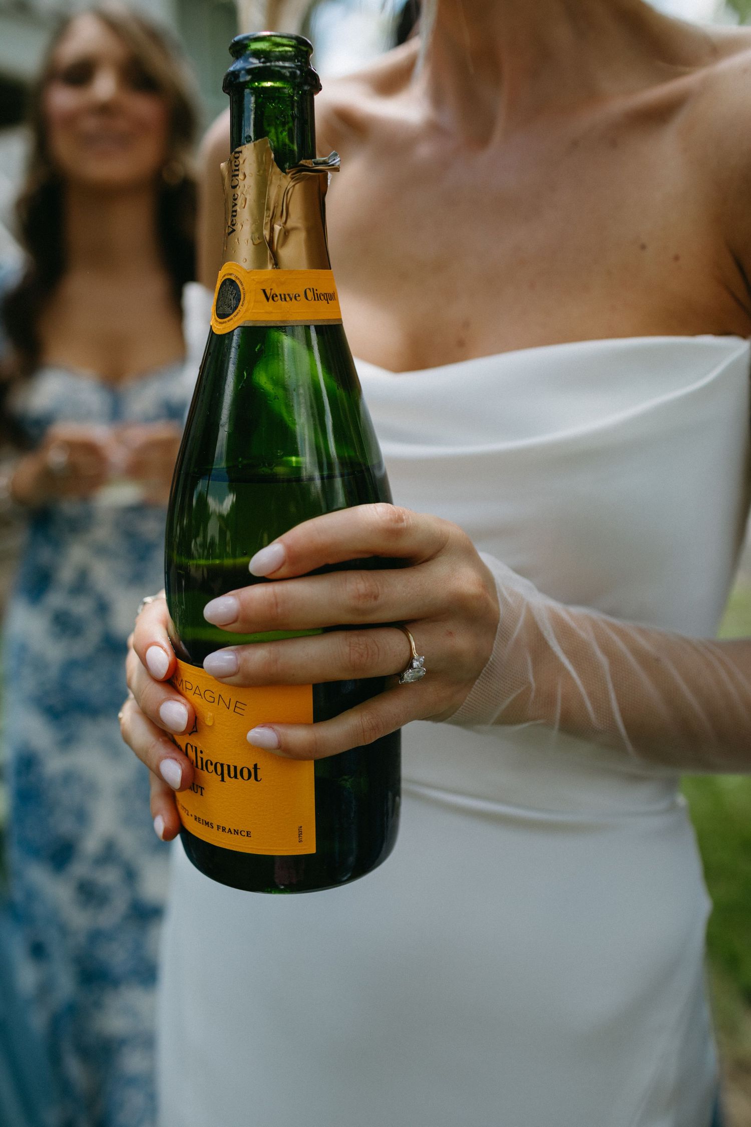 Bride in white gown holding Veuve Clicquot champagne bottle with engagement ring visible on her hand.