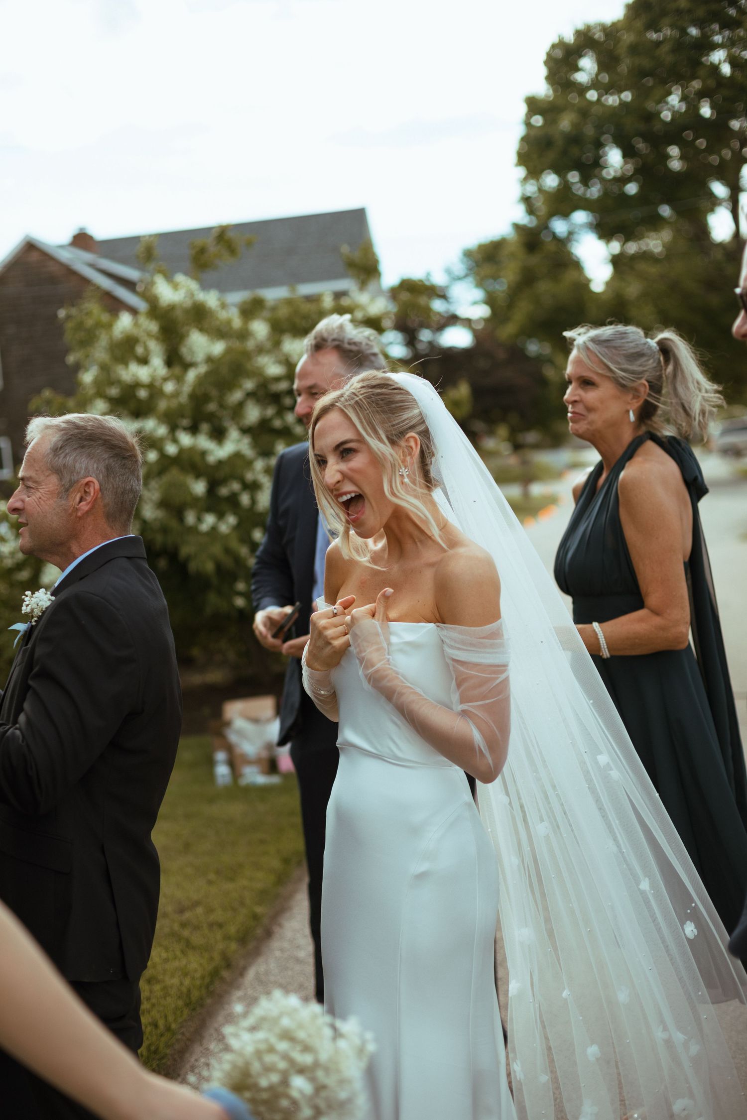 Excited bride in off-shoulder gown with star-dotted veil laughs joyfully during outdoor garden wedding ceremony.