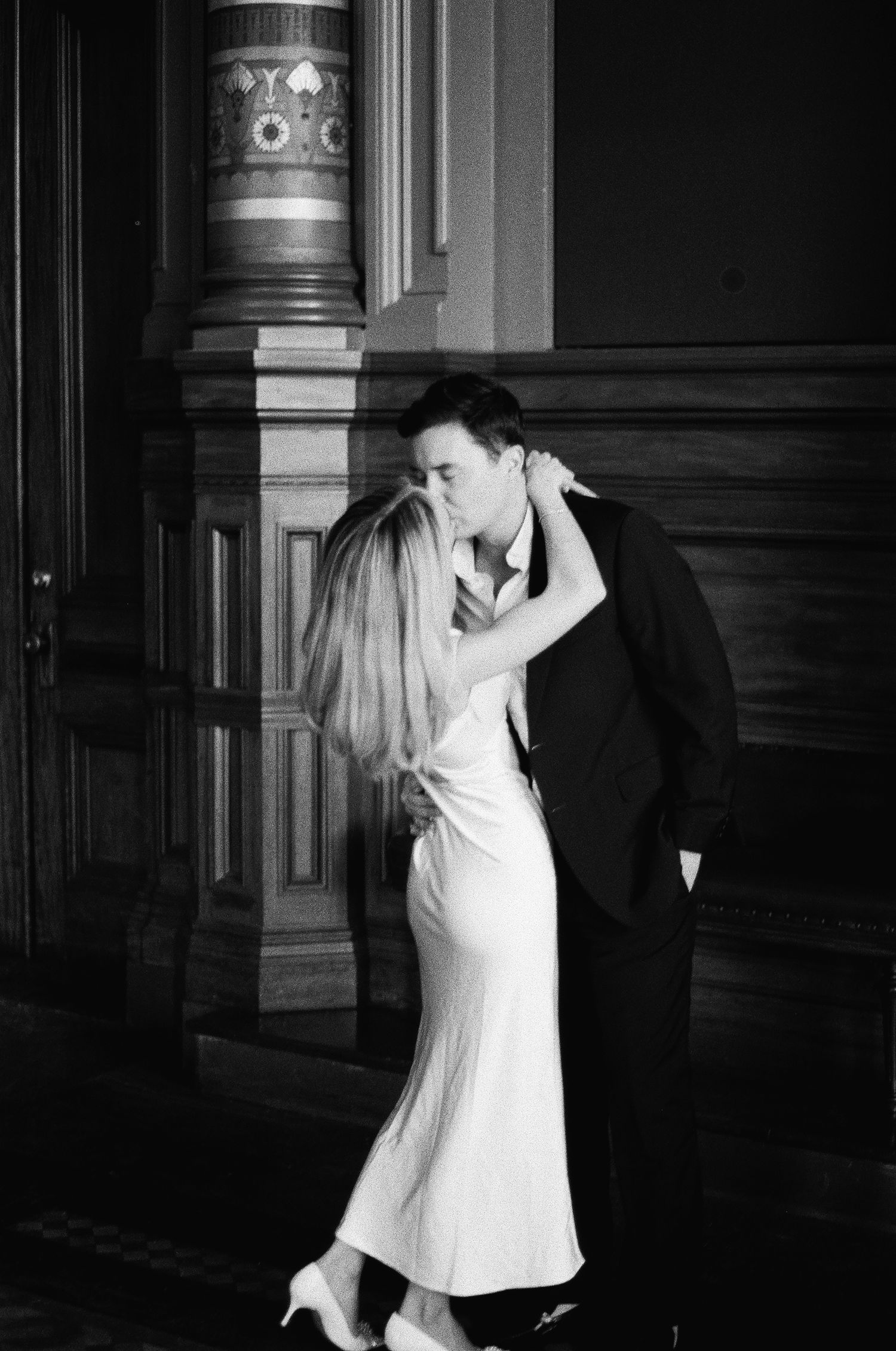 Newlyweds share romantic kiss beside ornate wooden column inside historic city hall building.