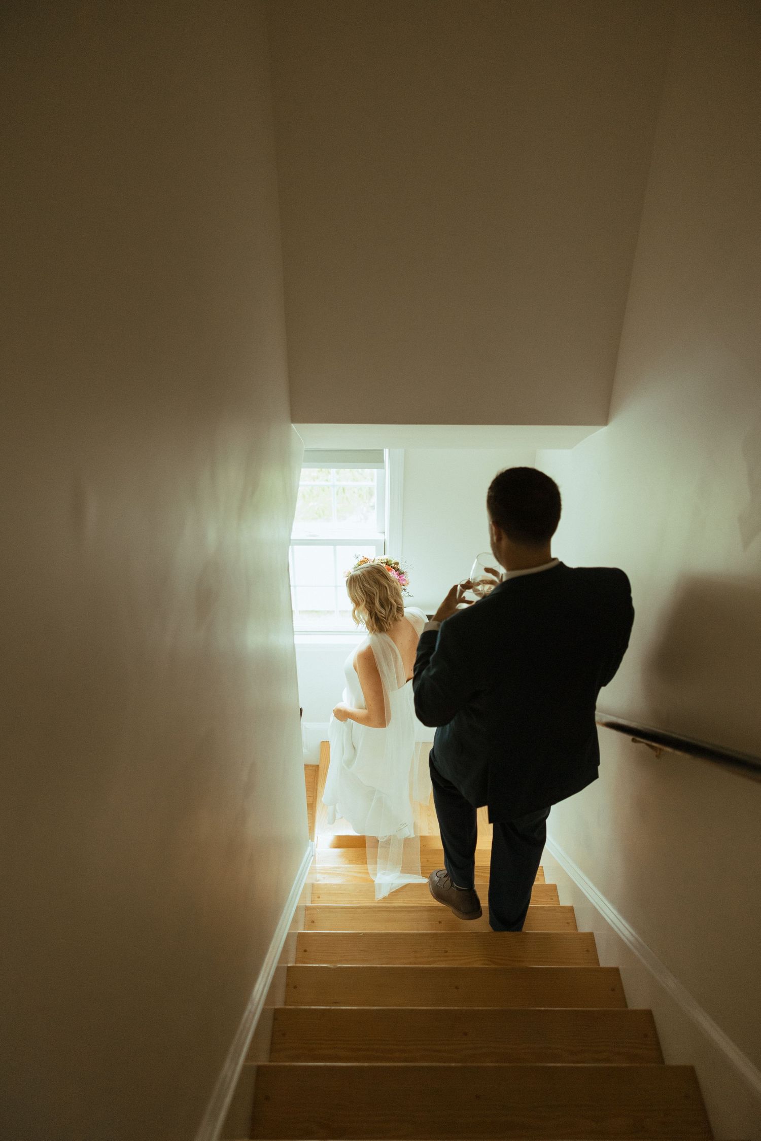 Groom photographs bride in white wedding dress at the bottom of indoor stairs near a bright window.