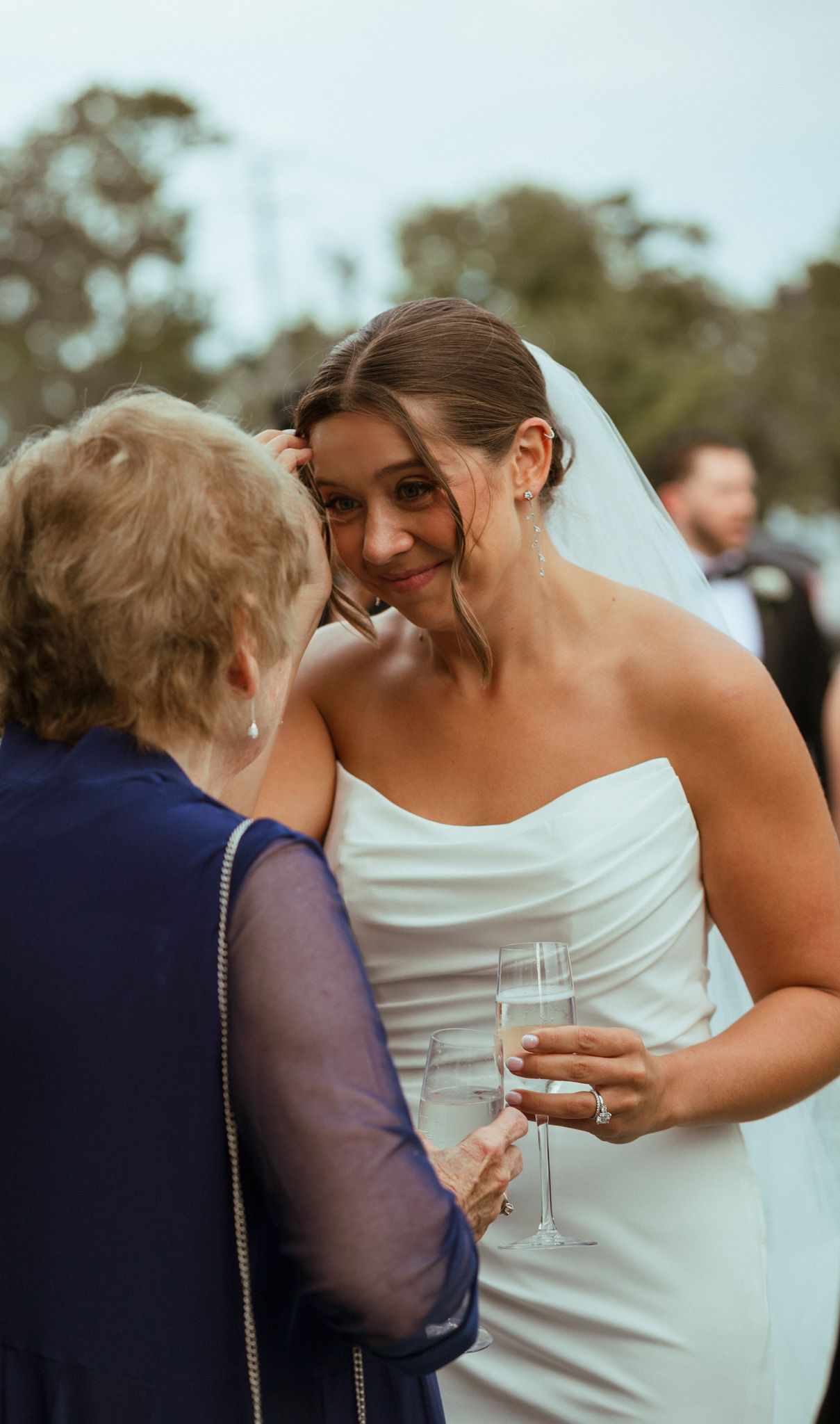 Smiling bride in white strapless gown holds champagne while sharing tender moment with elderly guest.