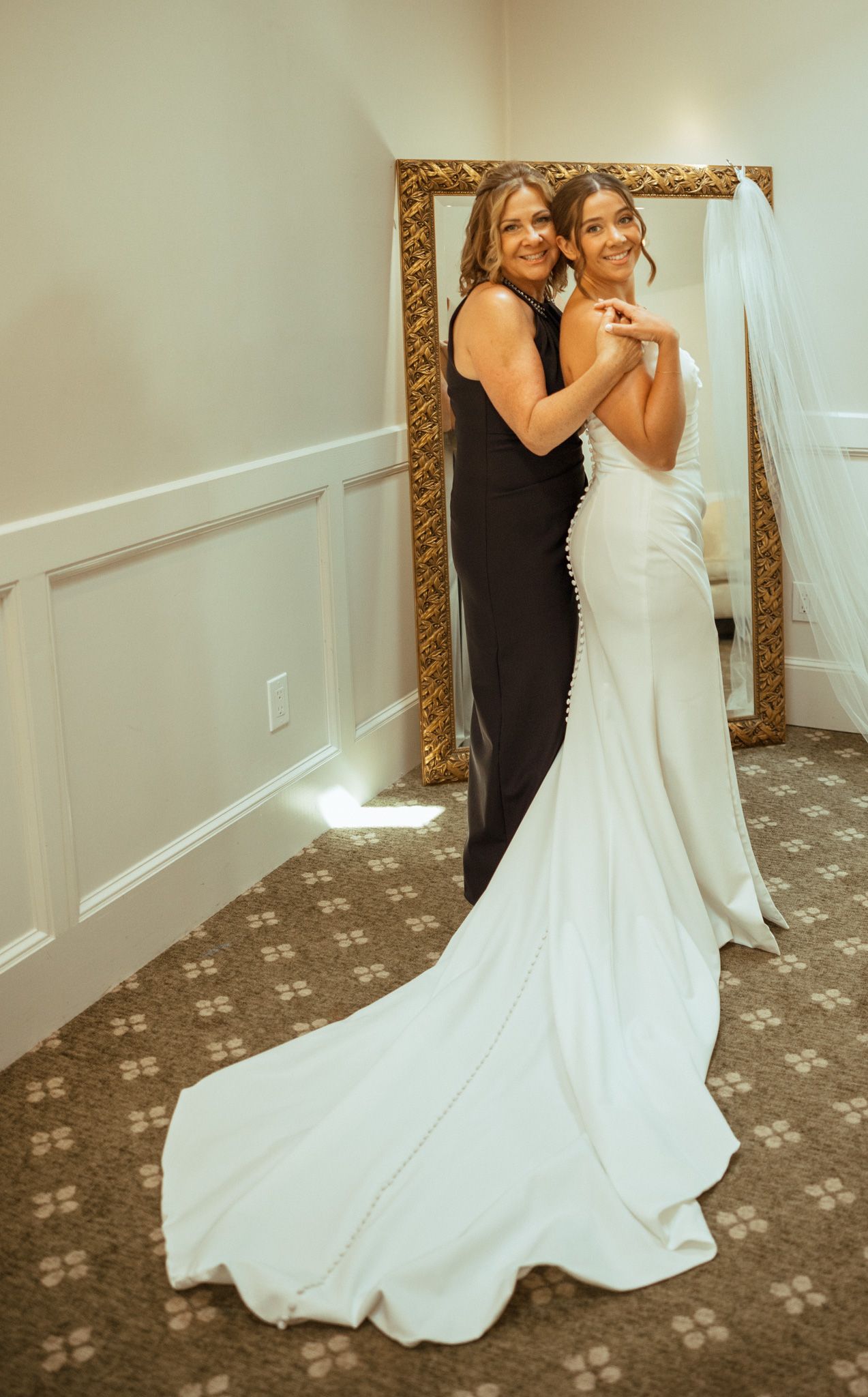 Bride in white mermaid gown posing with woman in black dress before gold mirror.