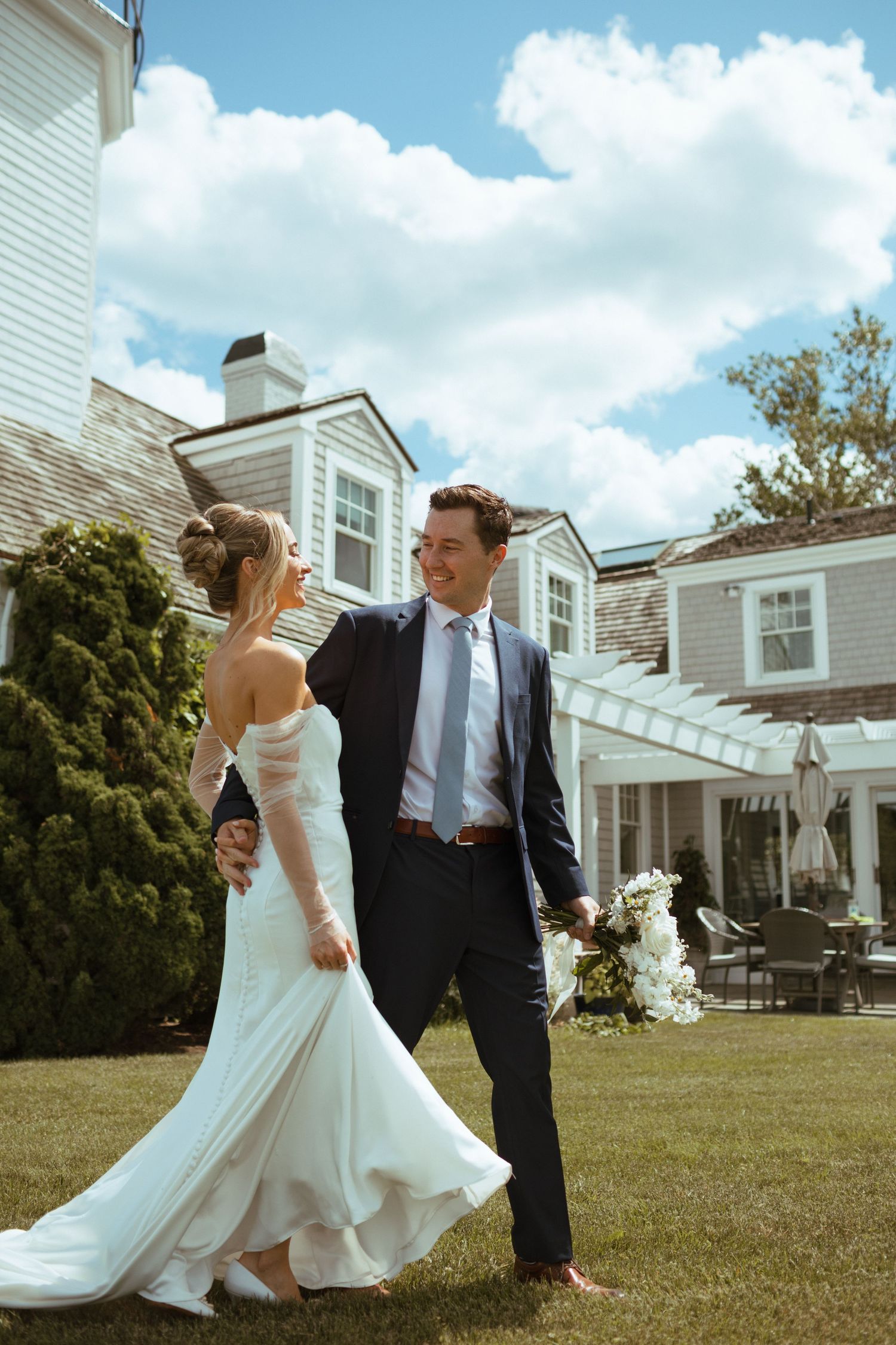 Bride and groom stroll across a sunny lawn, smiling at each other in front of a classic New England estate.