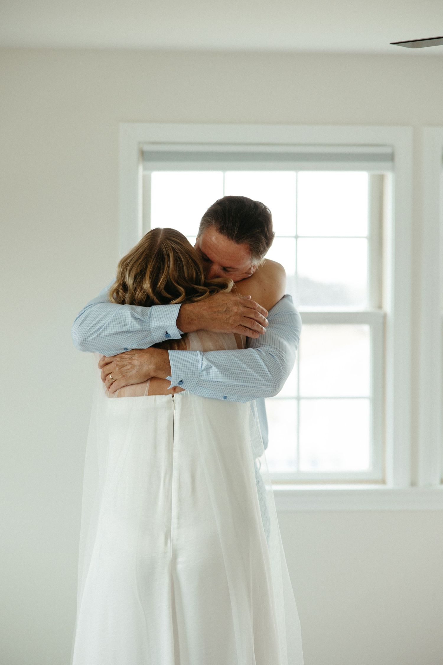Bride and father share a tender embrace in a bright white room before the wedding ceremony.