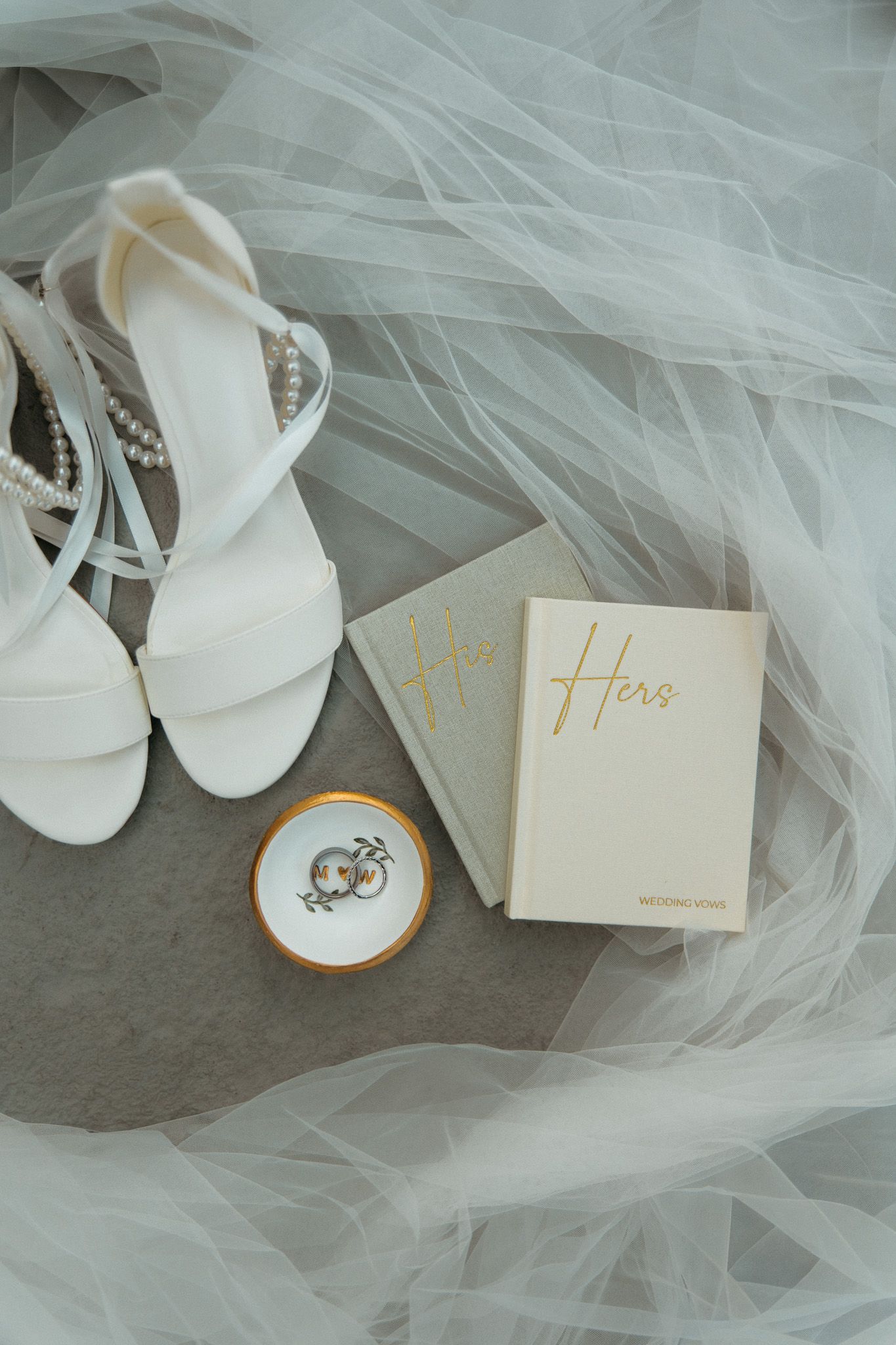 Flat lay of white bridal heels, wedding vow books, and ring dish on veil.