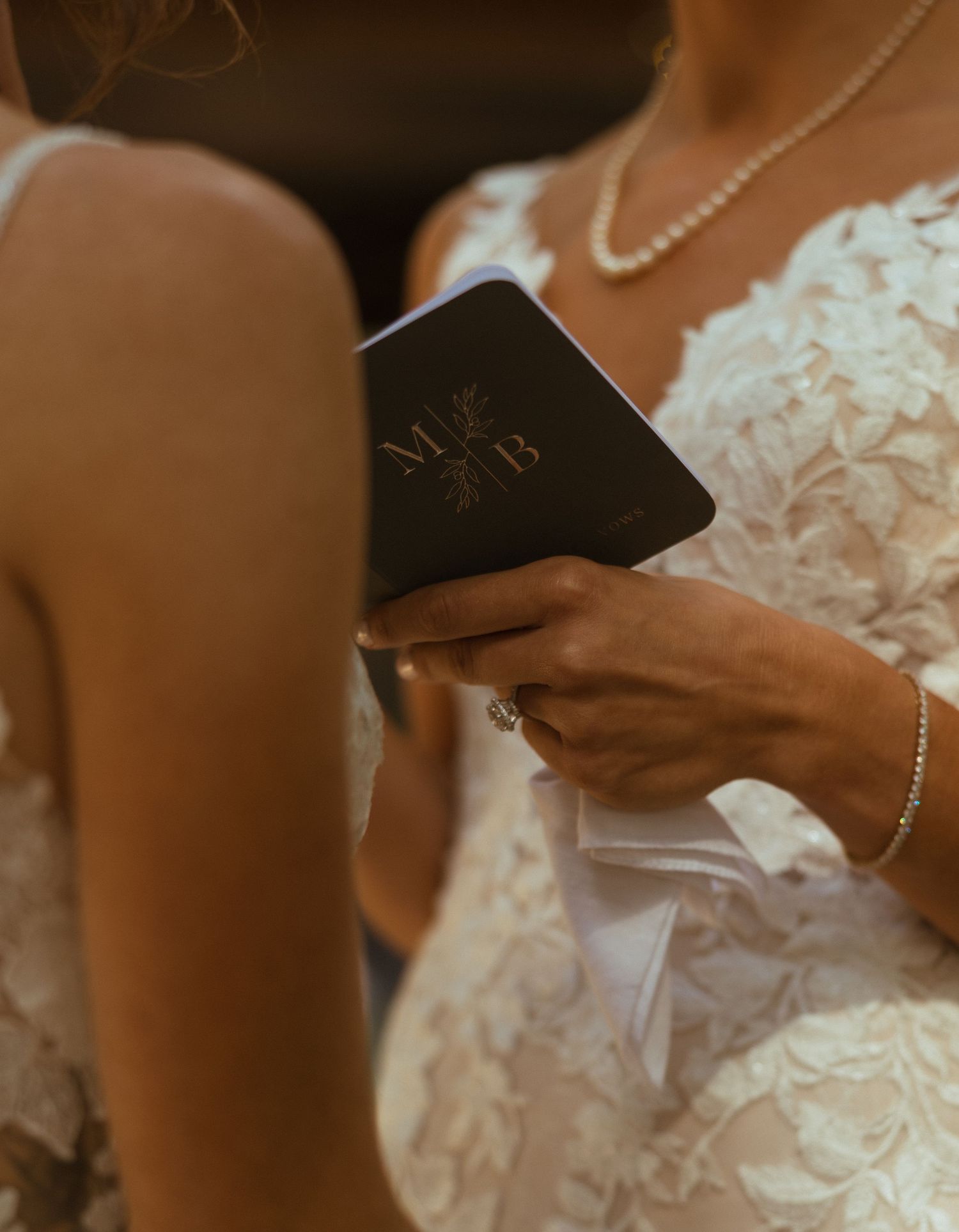 Close-up of bride holding monogrammed black vow booklet with initials M and B during wedding ceremony.