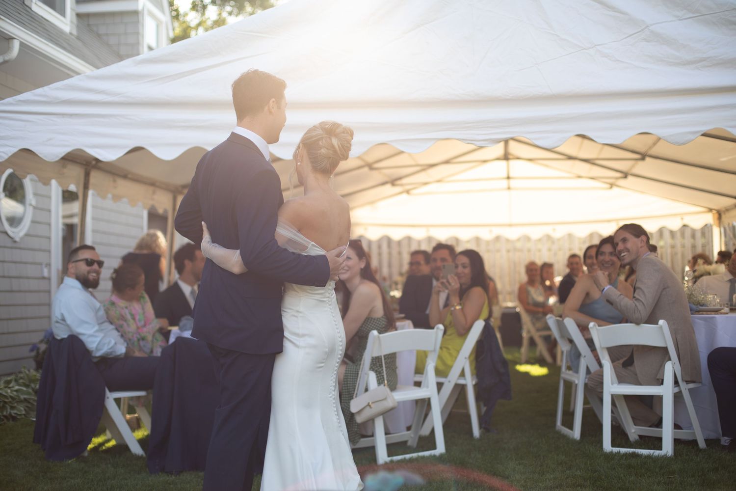 Bride and groom sharing first dance under white tent with seated wedding guests watching.