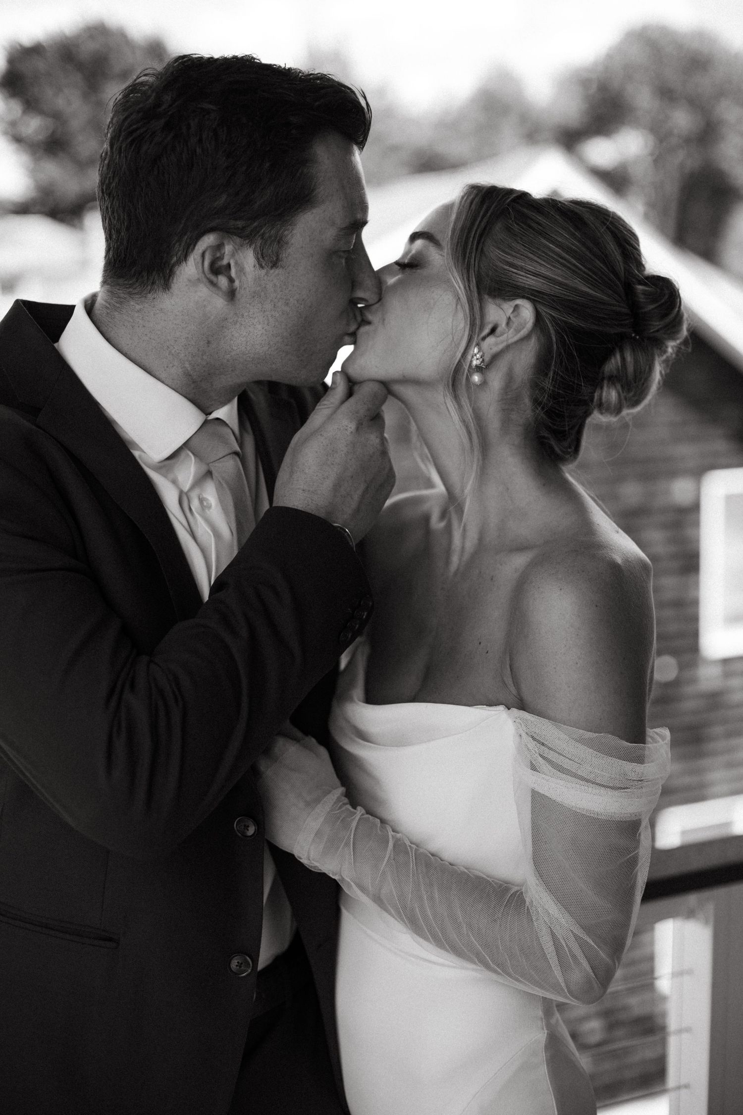 Black and white portrait of groom tenderly kissing bride in off-shoulder wedding gown outdoors.