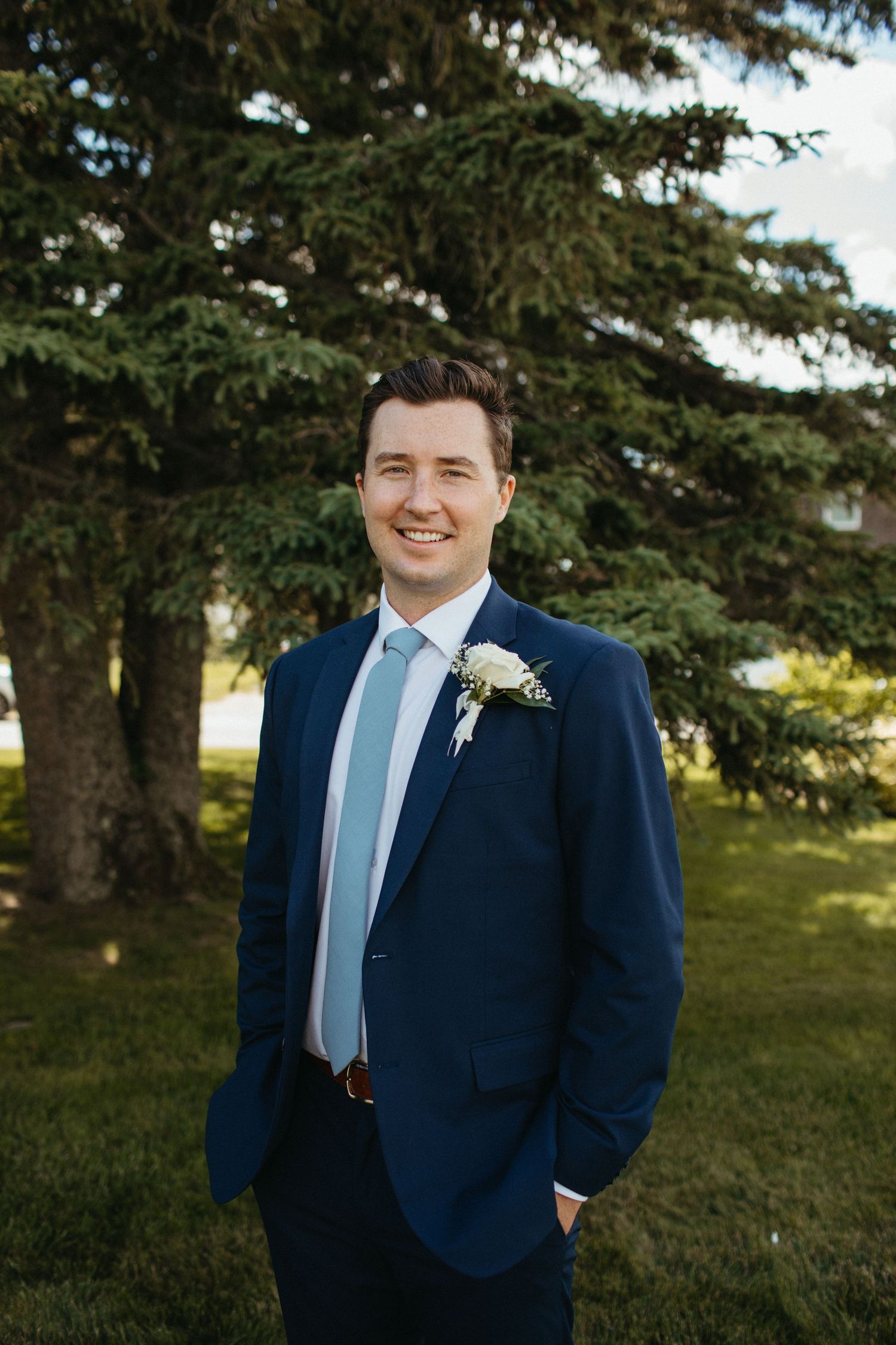Groom in navy blue suit with light blue tie and white boutonniere smiling outdoors near evergreen trees.
