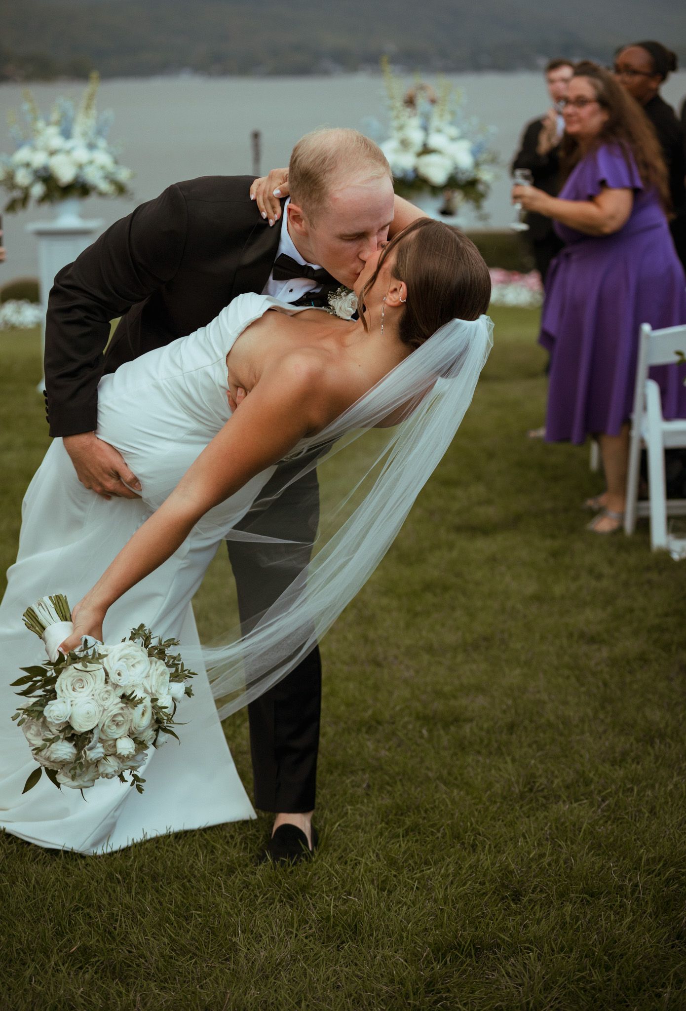 Groom dips bride for a romantic kiss during outdoor wedding recessional by the water.