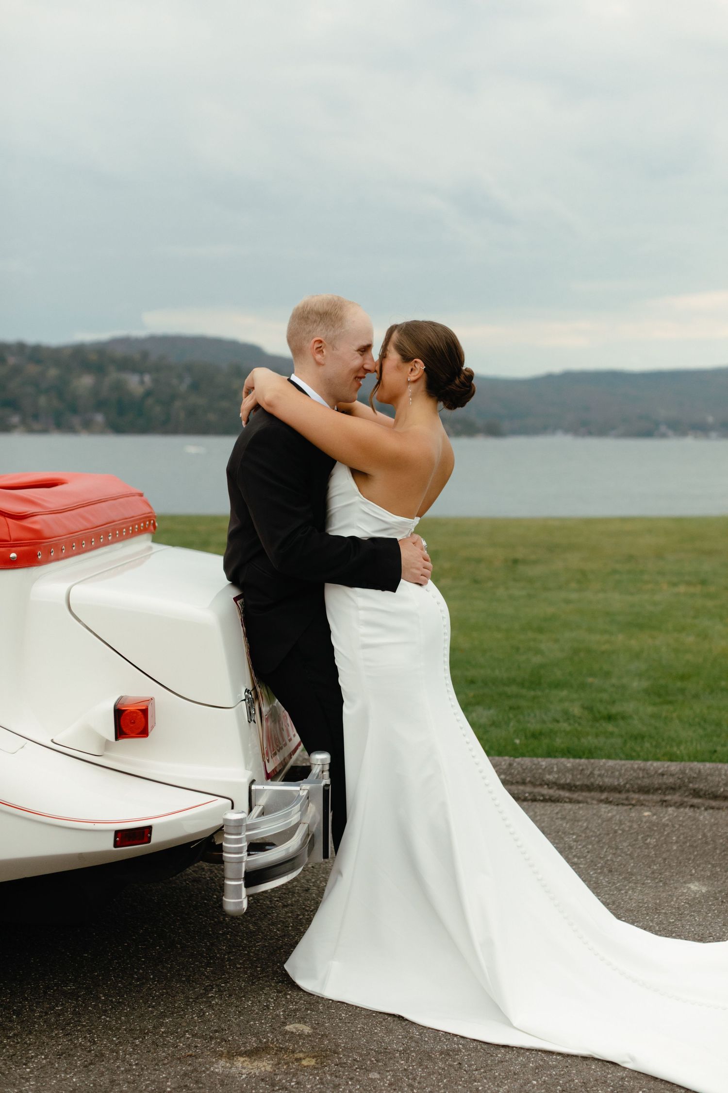 Newlyweds embracing and kissing beside a classic white convertible car with a scenic waterfront backdrop.