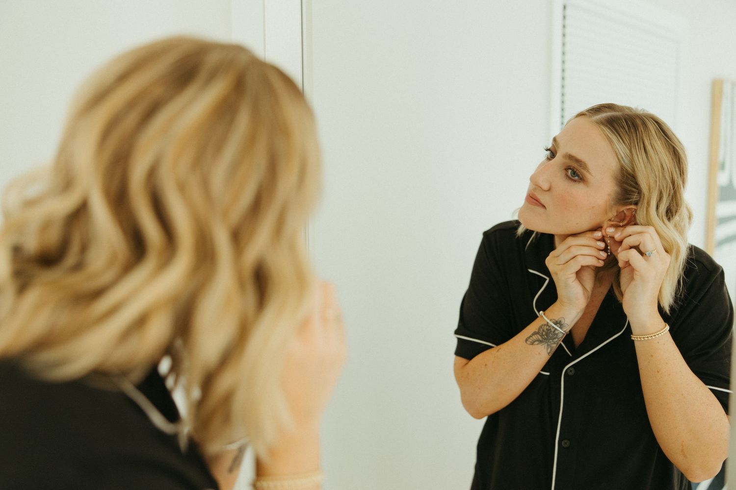 Blonde bride putting on earrings while looking in mirror during bridal preparations.