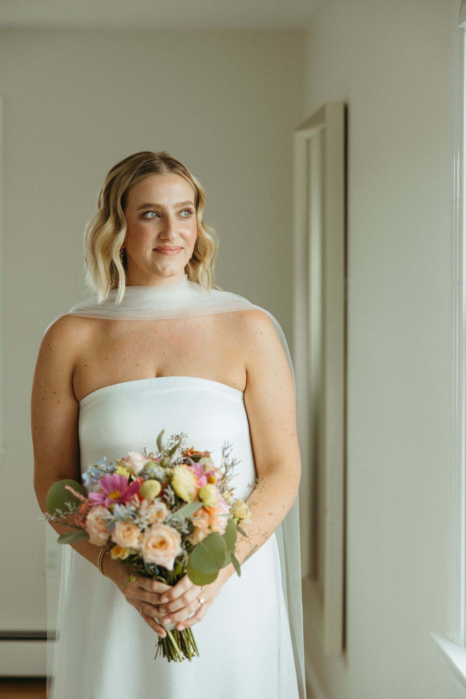 Smiling bride in strapless white gown holds colorful bouquet of pink, peach, and blue flowers.