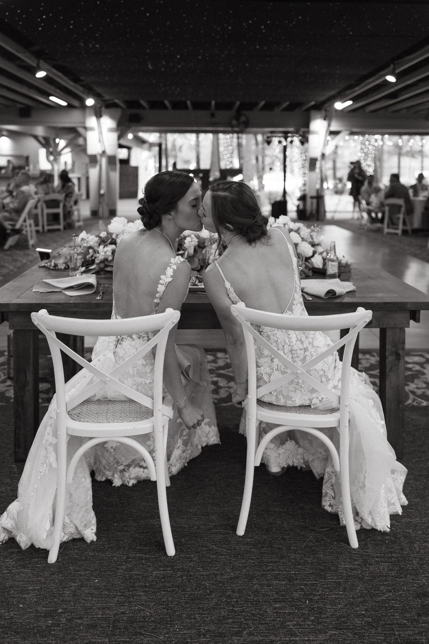 Black and white photo of two brides in lace gowns sitting close at sweetheart table.film-inspired