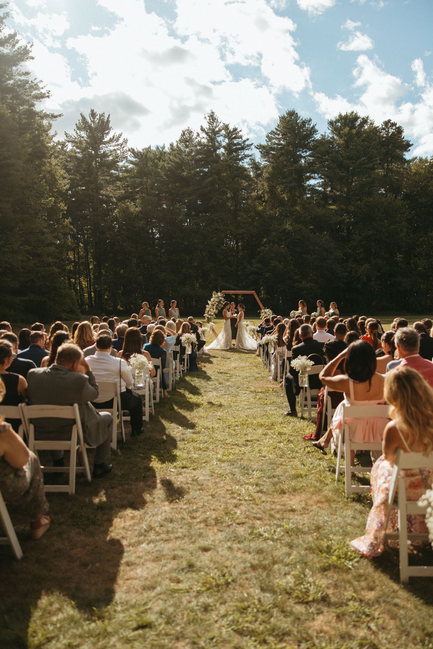 Wide view of outdoor wedding ceremony with guests seated in white chairs facing floral arch altar.