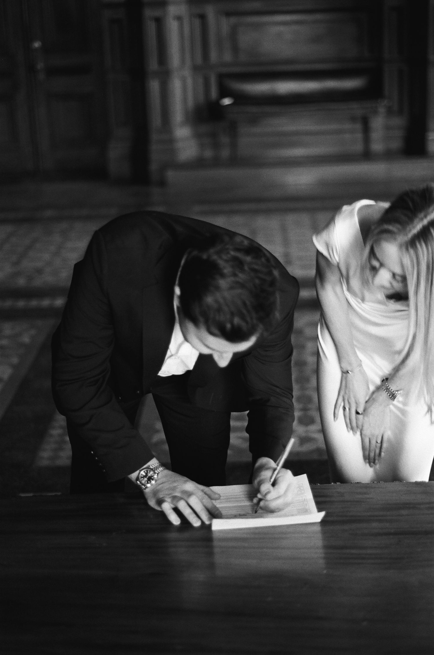 Groom in black suit signs wedding documents at wooden table while bride in white dress looks on.film-inspired