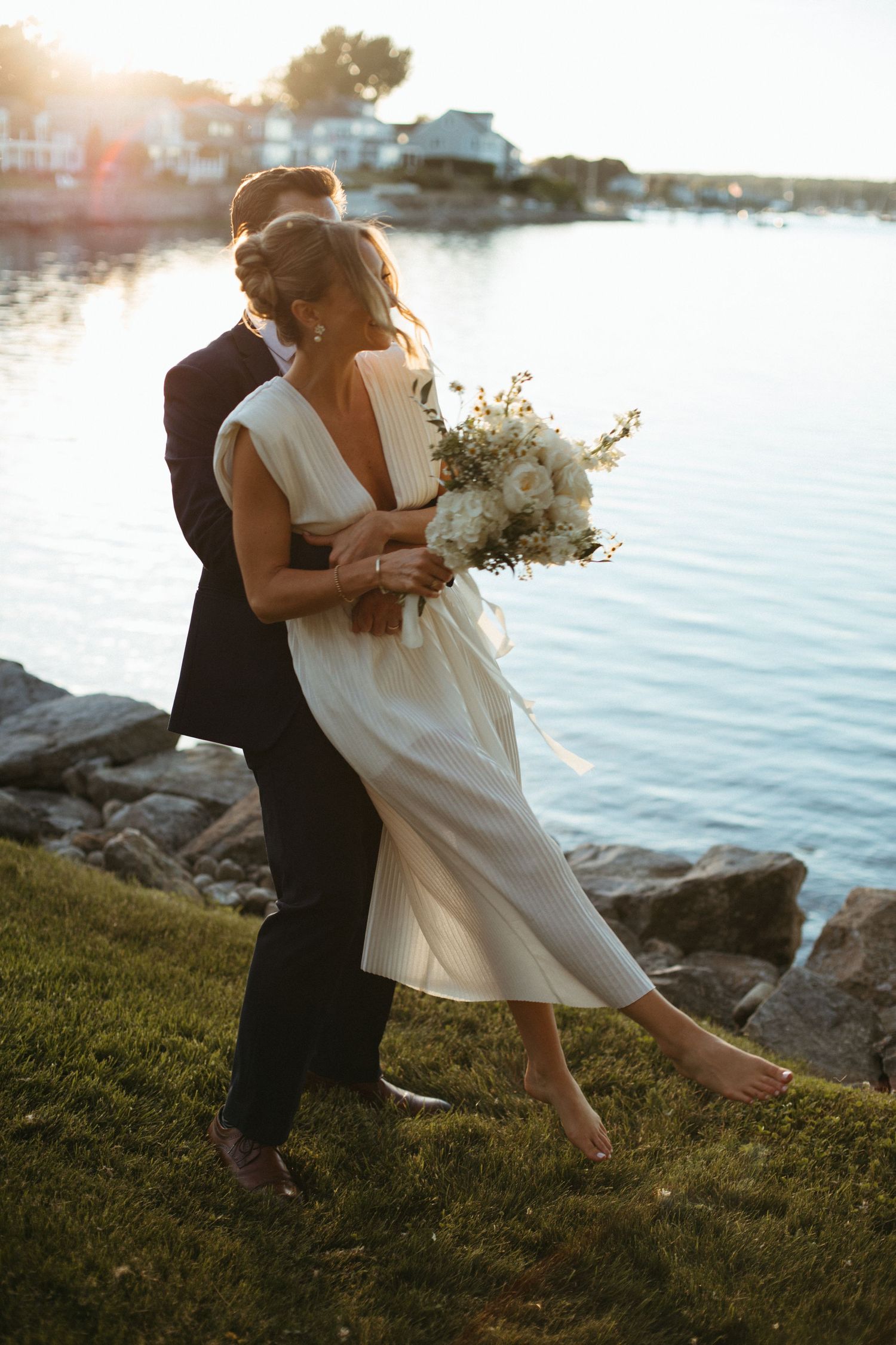 Groom embracing bride holding white bouquet by rocky waterfront during stunning golden hour sunset.