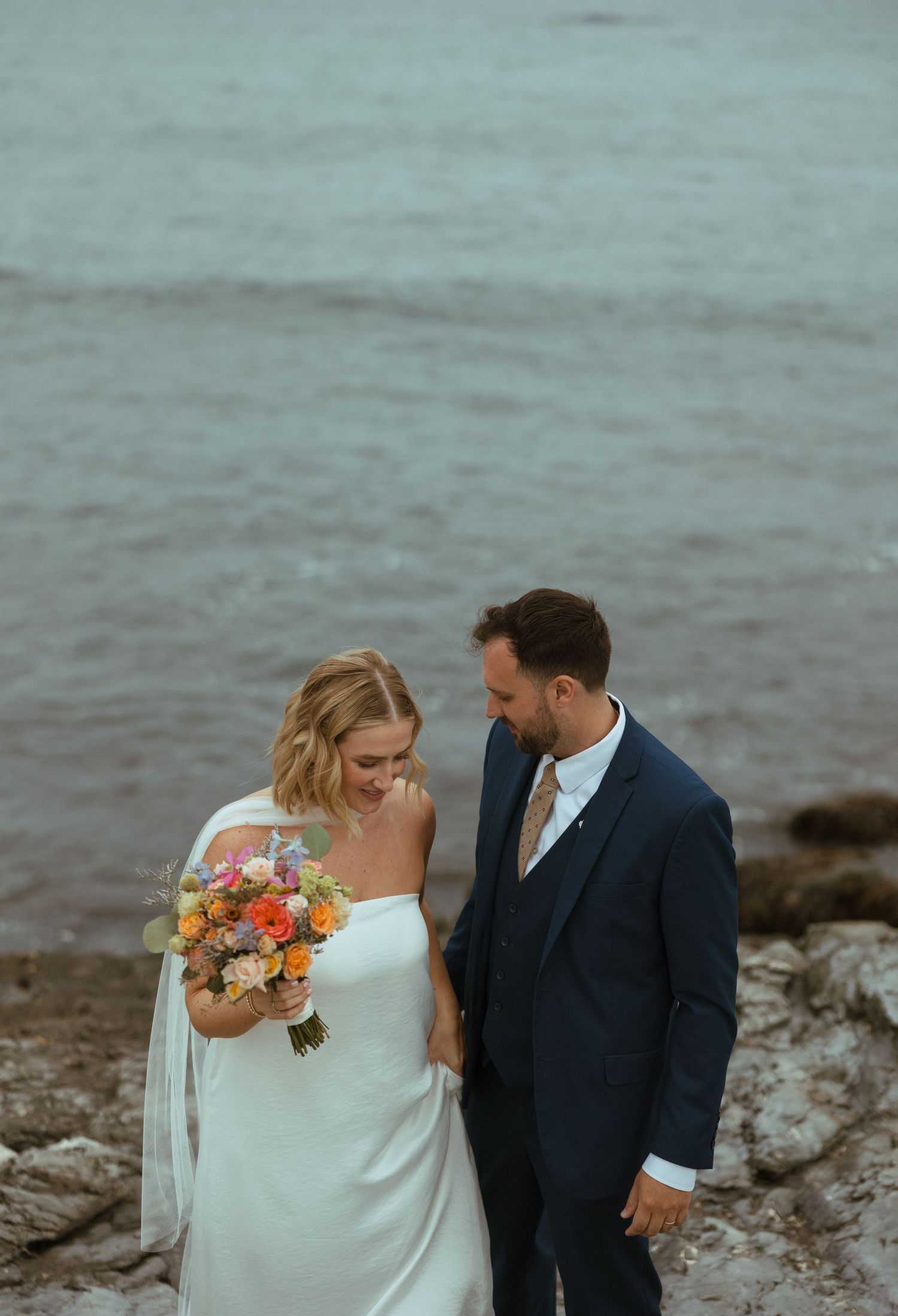 Bride laughs joyfully holding a bright colorful bouquet beside her smiling groom on a rocky oceanside setting.