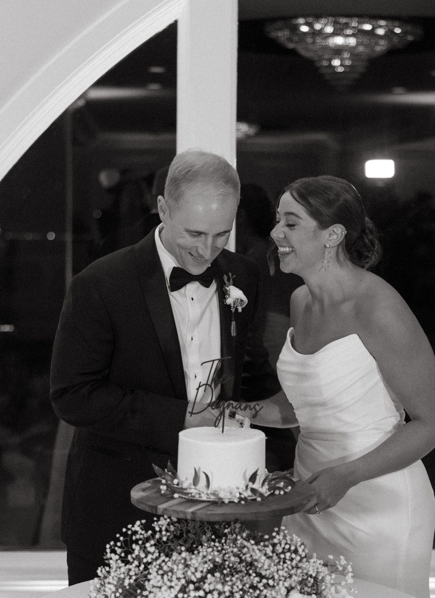 Black and white photo of bride and groom laughing while cutting wedding cake.