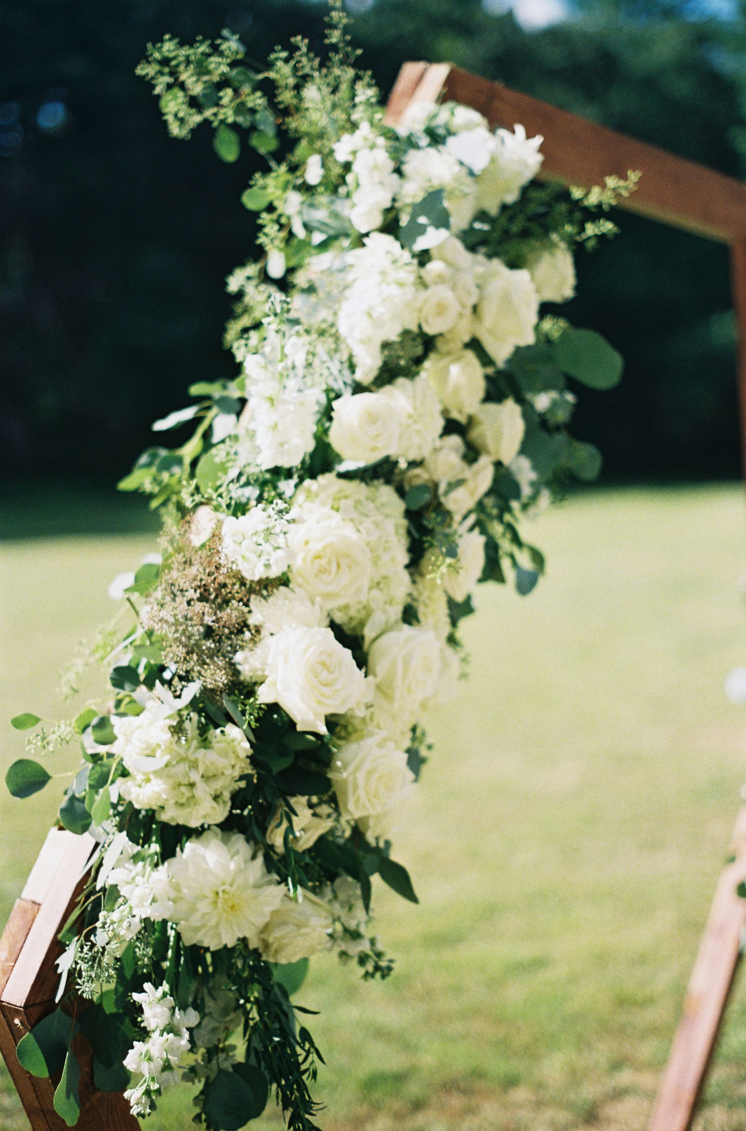 Elegant white floral arrangement with roses and greenery adorning wooden wedding arch.film-inspired