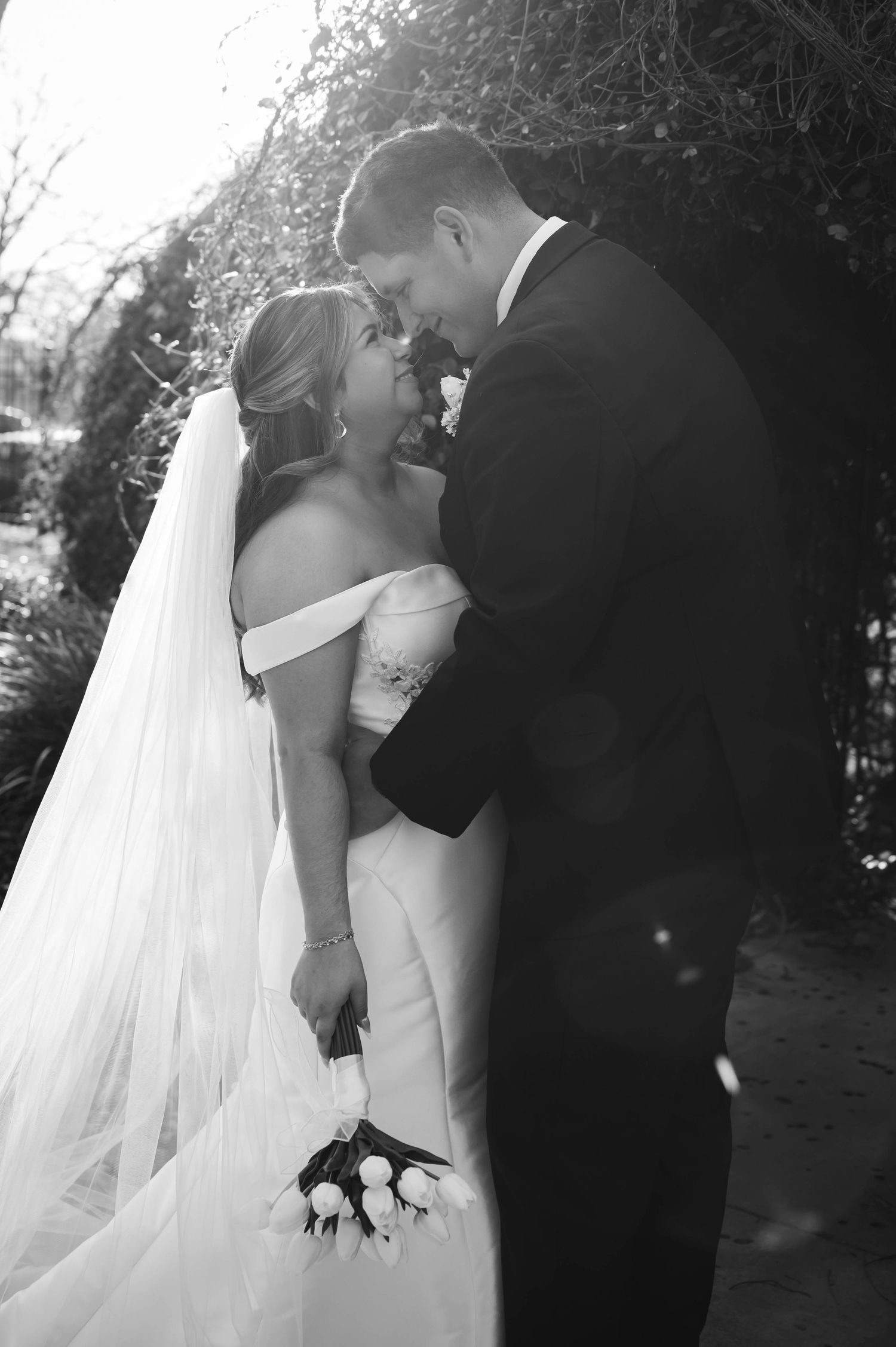 Black and white photo of a bride and groom sharing an intimate moment, bride holding roses in an off-shoulder gown with veil.