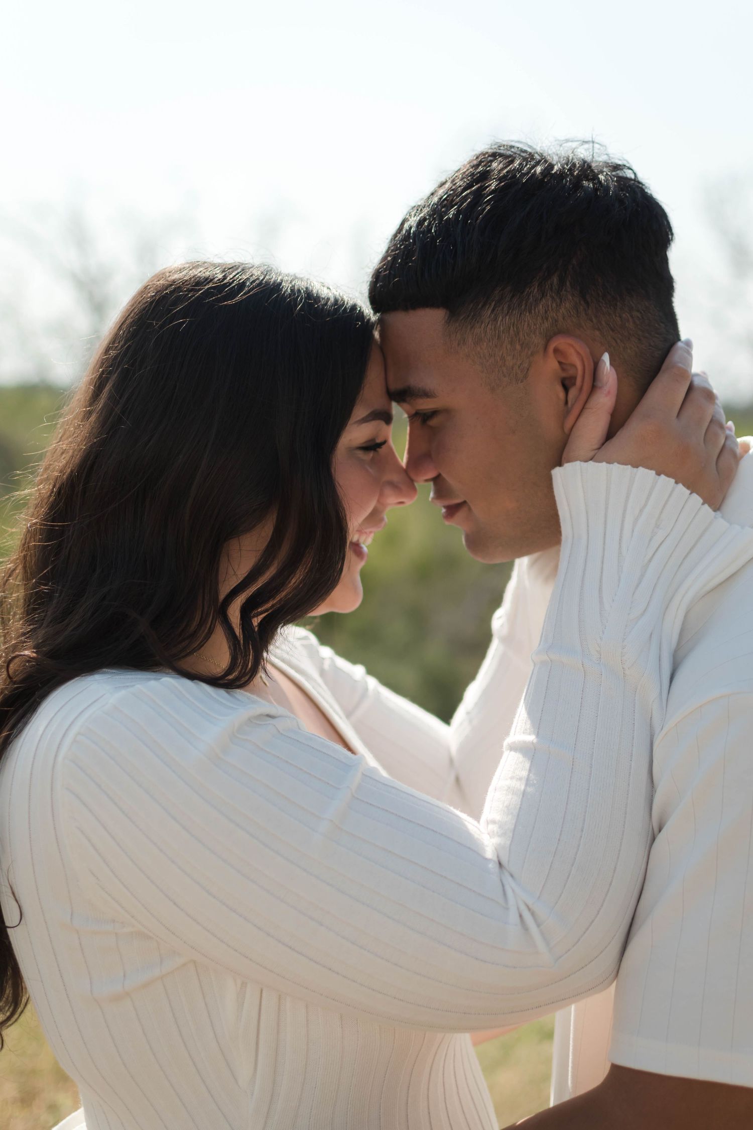 A romantic couple in white sweaters touching foreheads outdoors, sharing an intimate moment in natural sunlight.