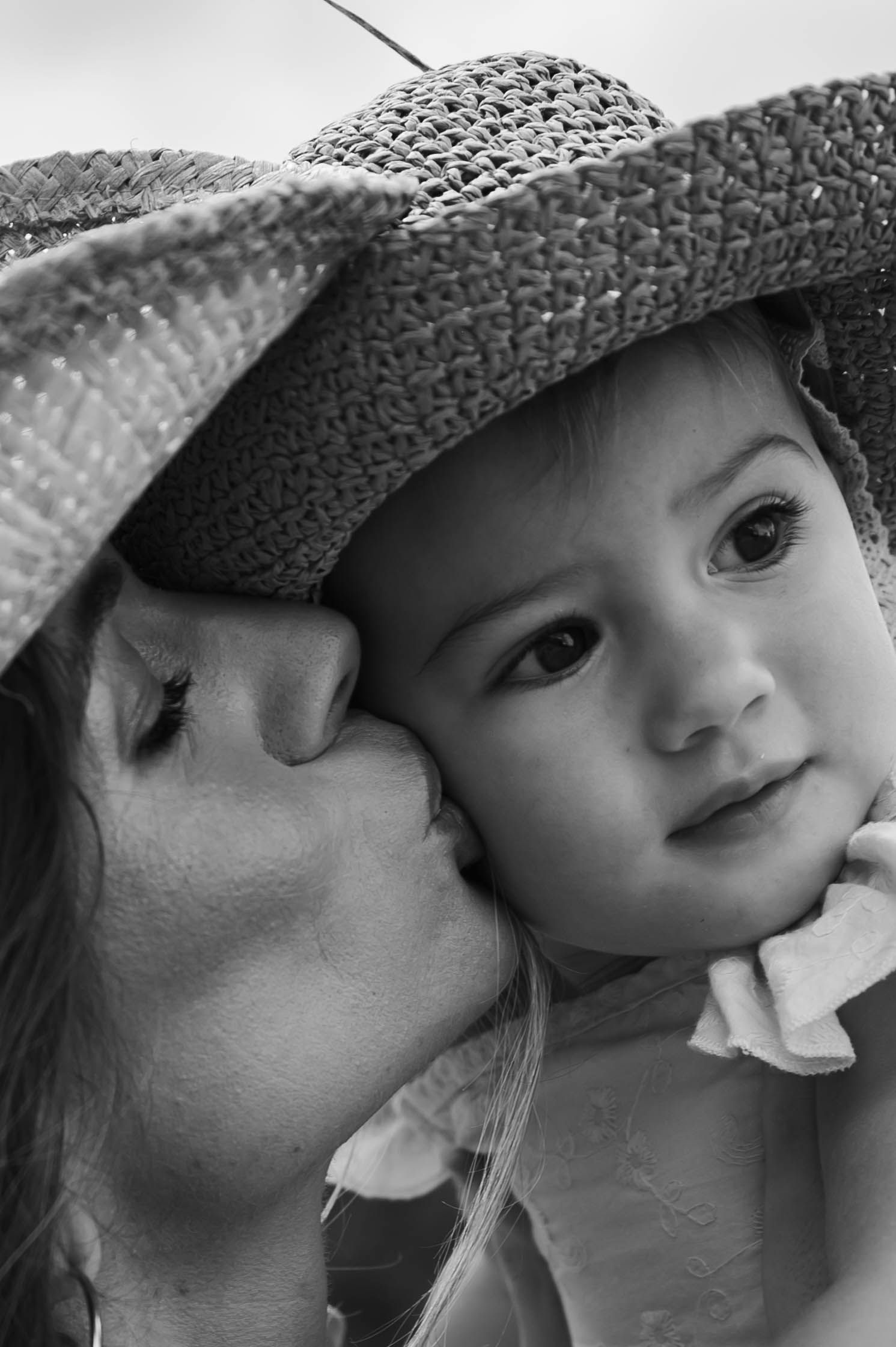 Black and white photo of a mother kissing her young child on the cheek, both wearing a wide-brim straw hat.