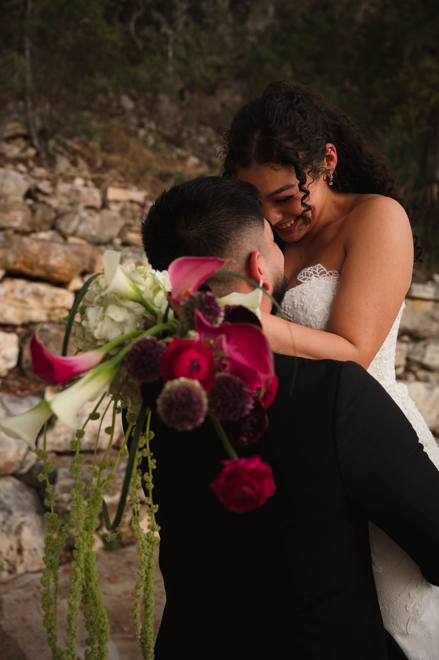 Groom lifts bride in wedding dress near stone wall as she smiles holding pink floral bouquet.