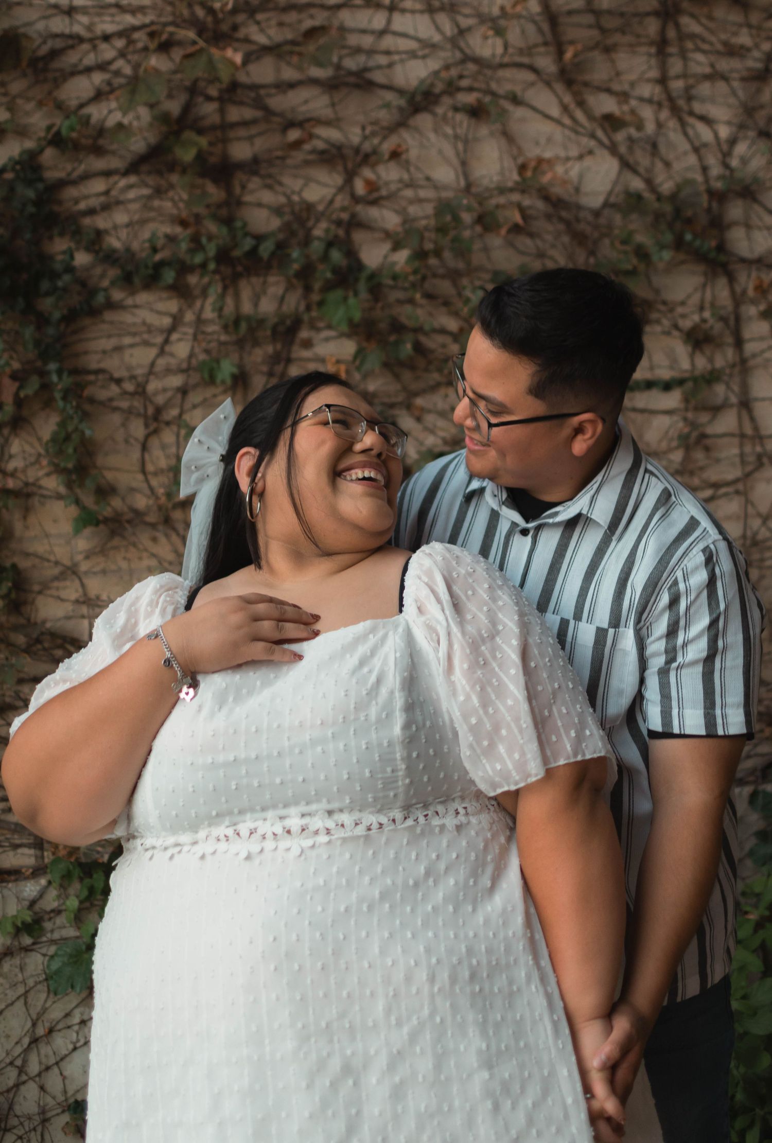 Happy couple in white dress and striped shirt laughing together against ivy-covered wall.