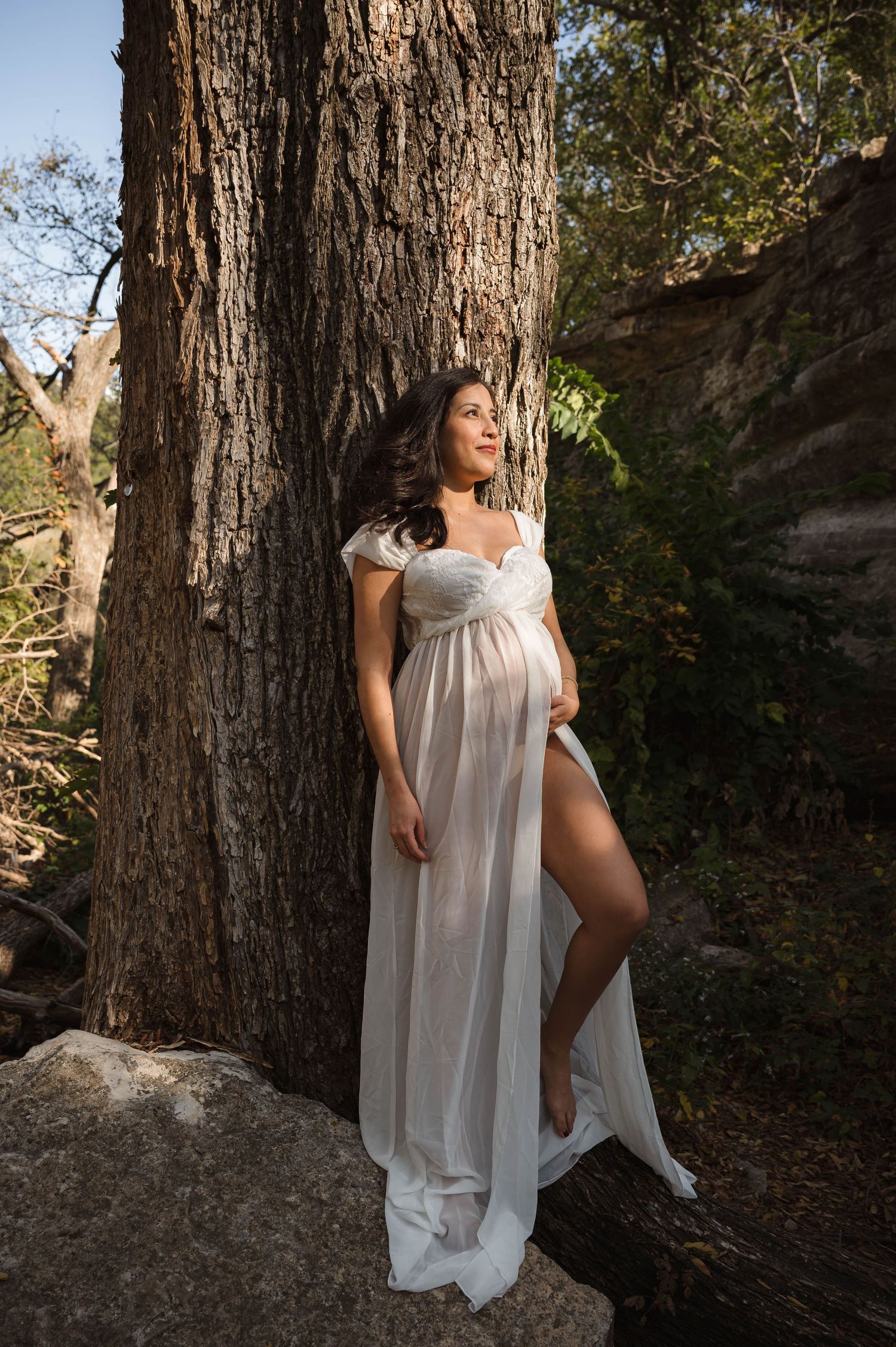 Pregnant woman in white sheer gown leaning against a tree in a forest, cradling her baby bump in natural light.
