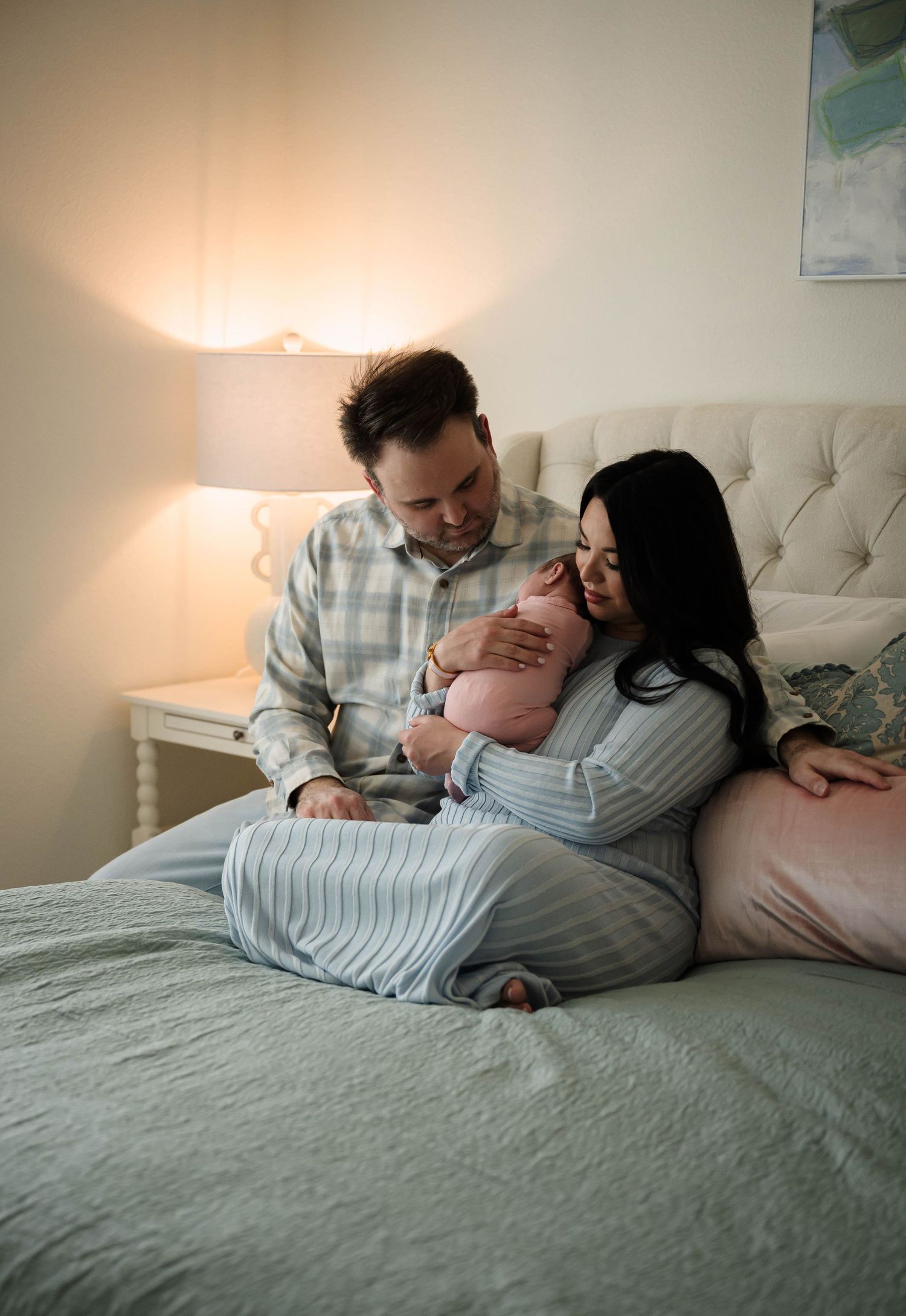 New parents sitting on bed gazing lovingly at their newborn baby wrapped in pink, soft bedroom lighting.