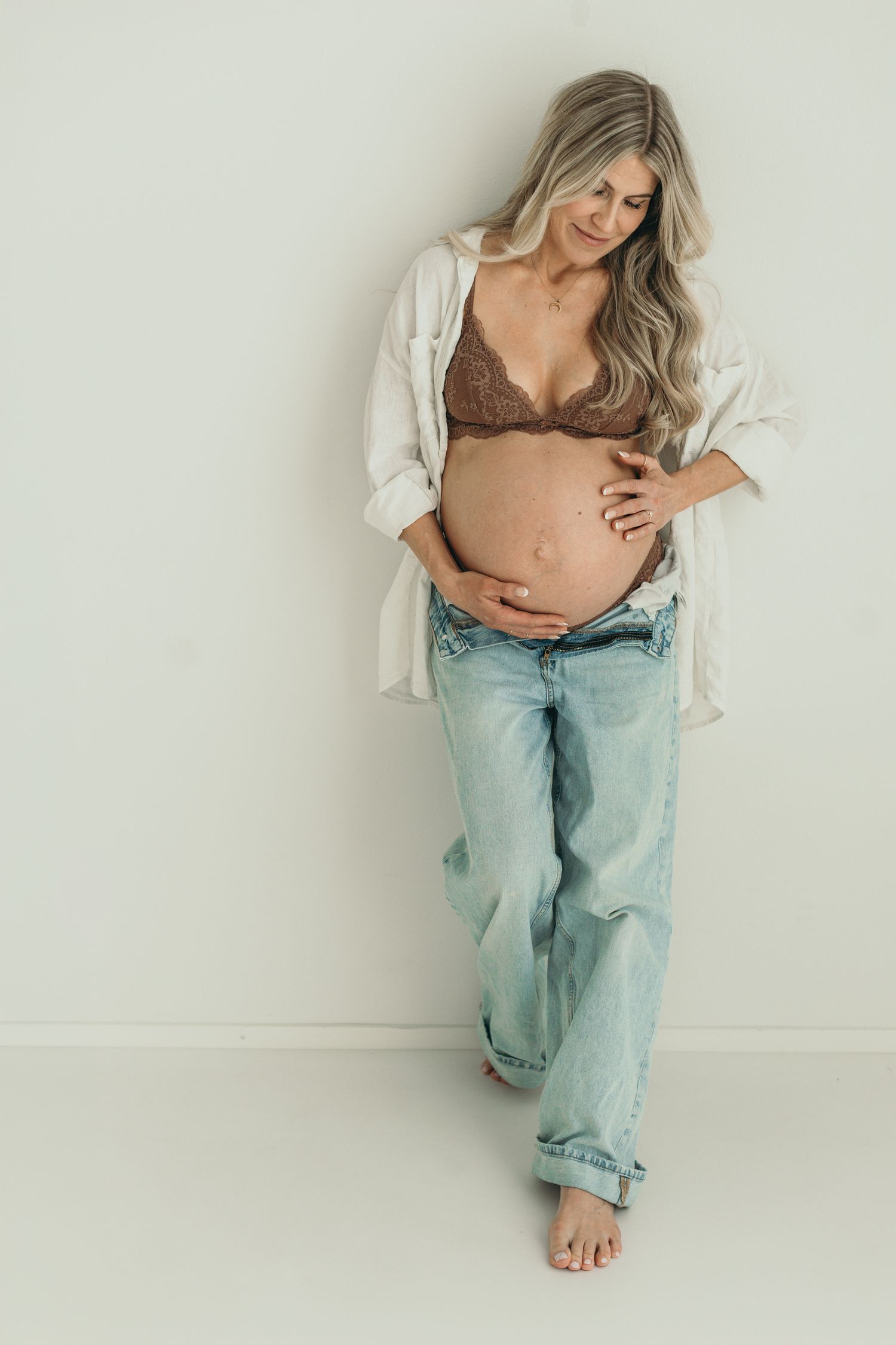 Pregnant woman in lace bra, open white shirt, and jeans cradling baby bump against light background.