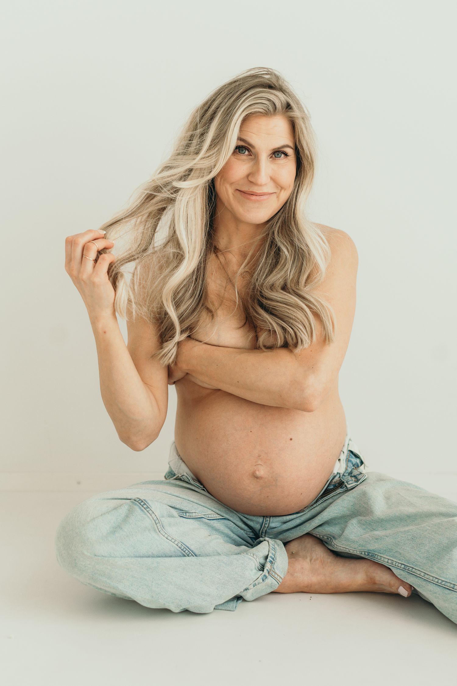 Pregnant woman with long blonde hair sitting cross-legged topless in jeans on white background. Maternity photography at the studio.