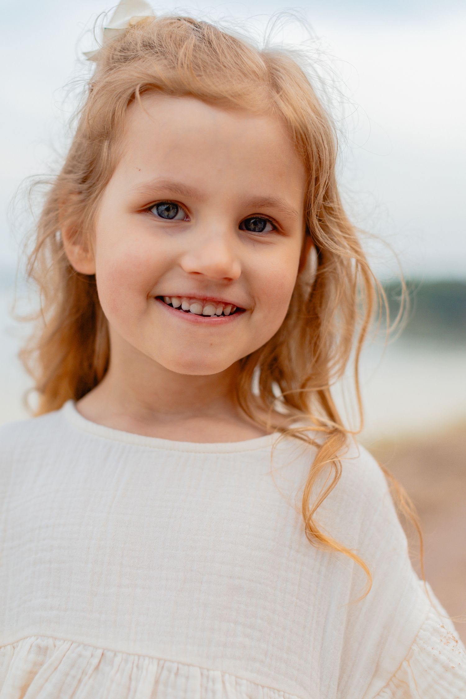 Smiling young girl with curly blonde hair and blue eyes wearing white dress at the beach. Lapsikuvaus miljöössä.